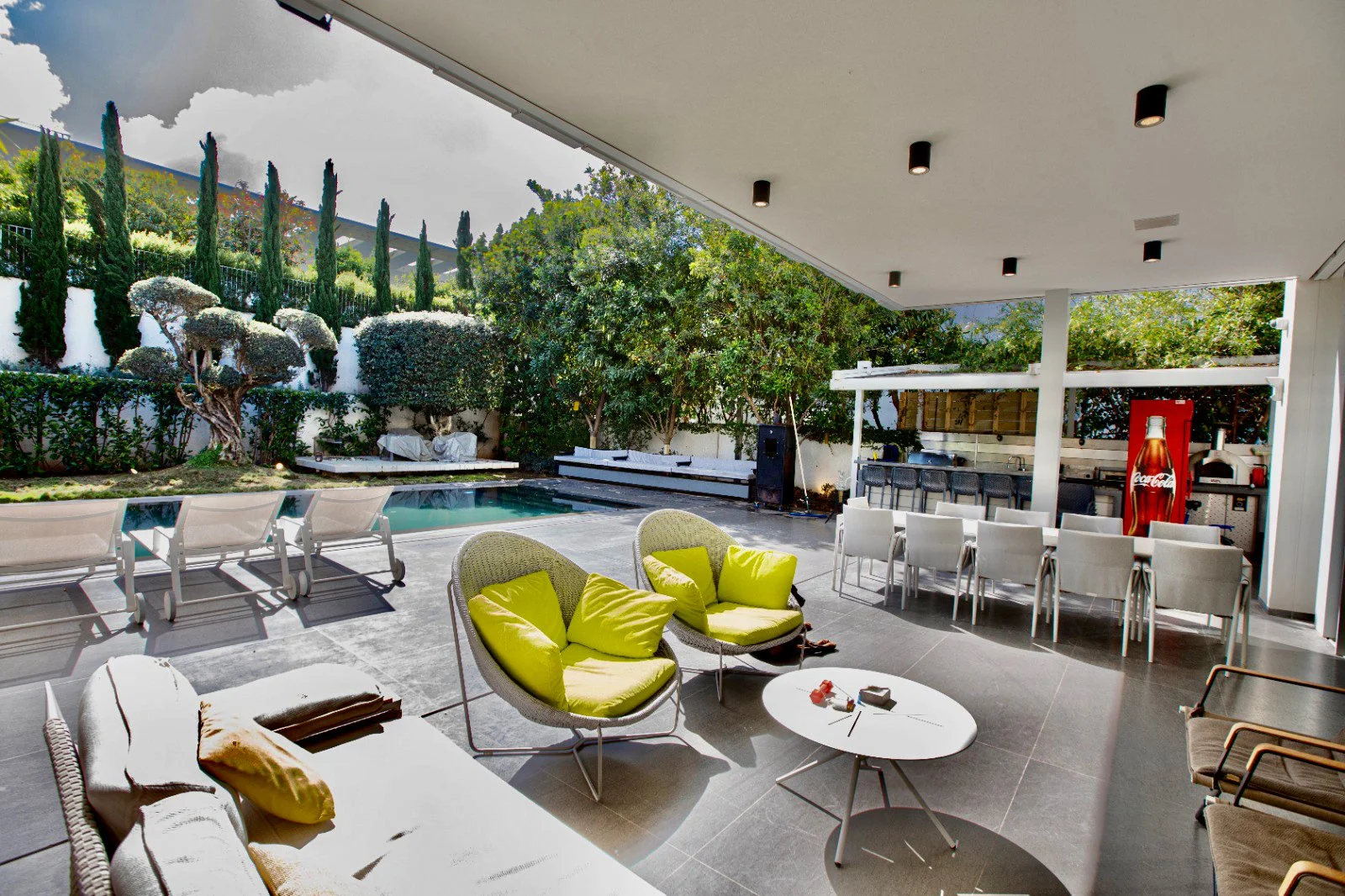 Covered outdoor patio with a seating area, a pool, and a bar with chairs and a Coca-Cola vending machine.