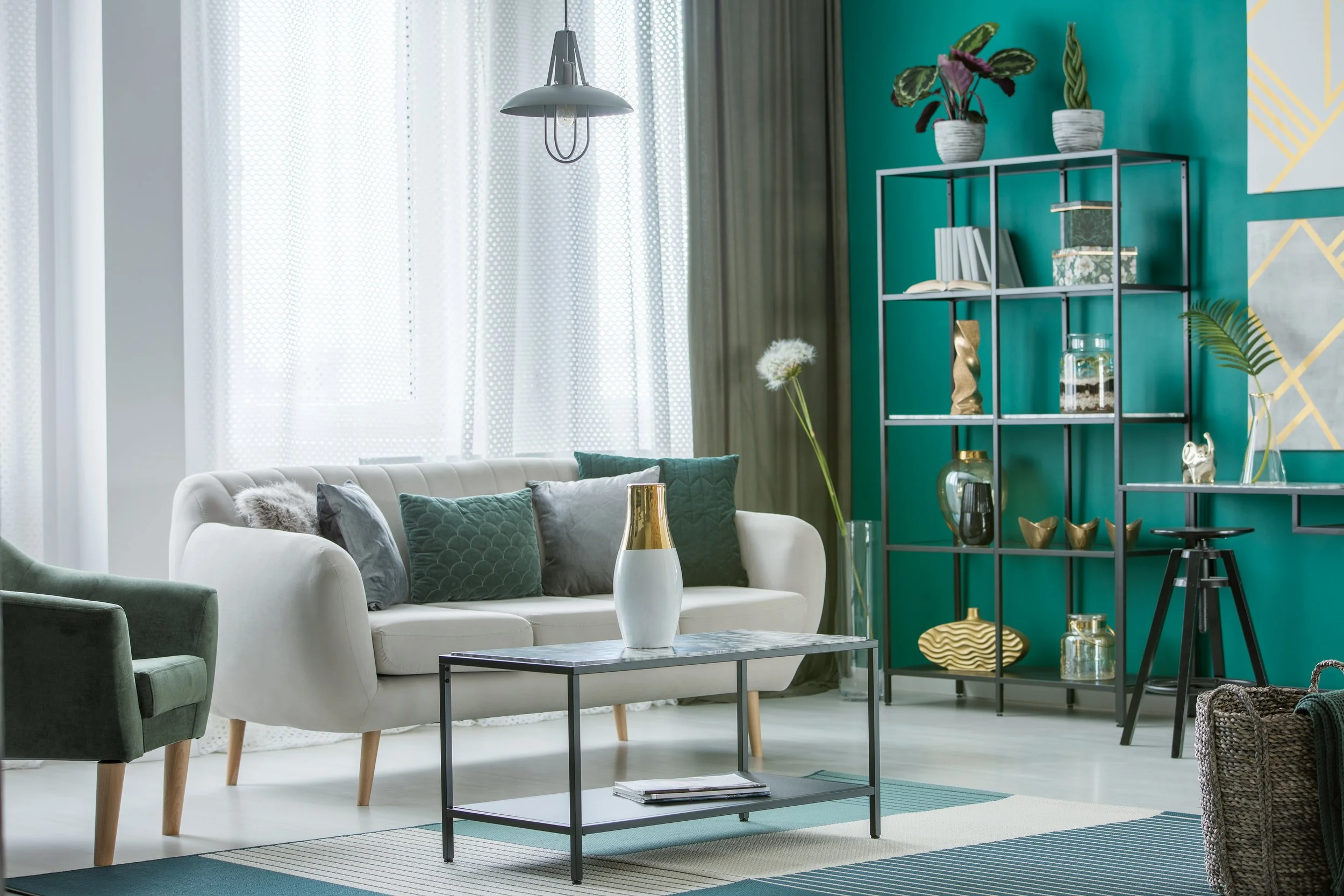Living room with white sofa, green armchair, teal accent wall, black metal bookshelf with decor, glass vases, and plants, sitting area with coffee table and striped rug, sheer curtains, and pendant light.