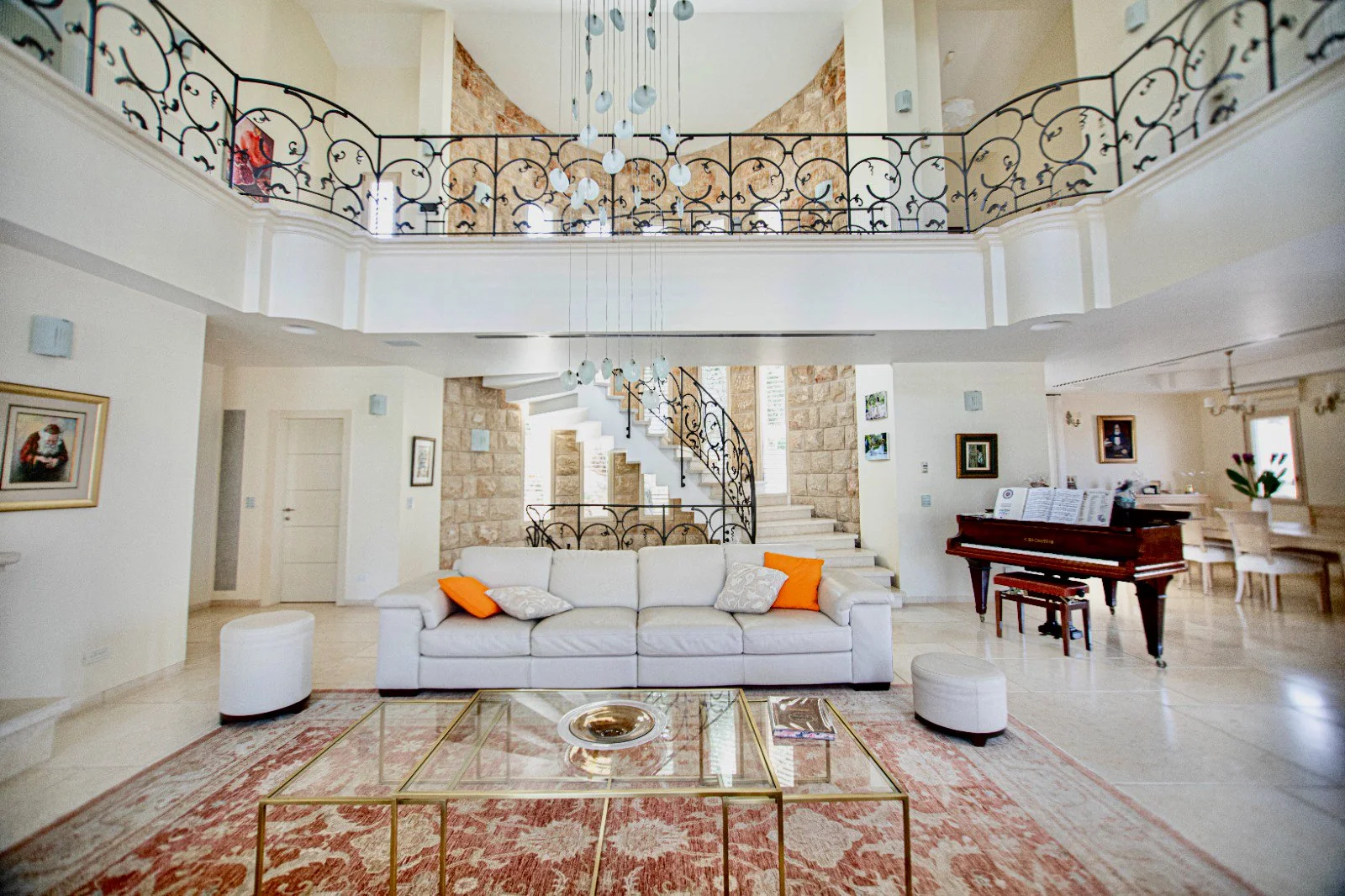 Bright living room with white sofa, orange throw pillows, glass coffee table, grand staircase, chandelier, grand piano, and decorative railings.