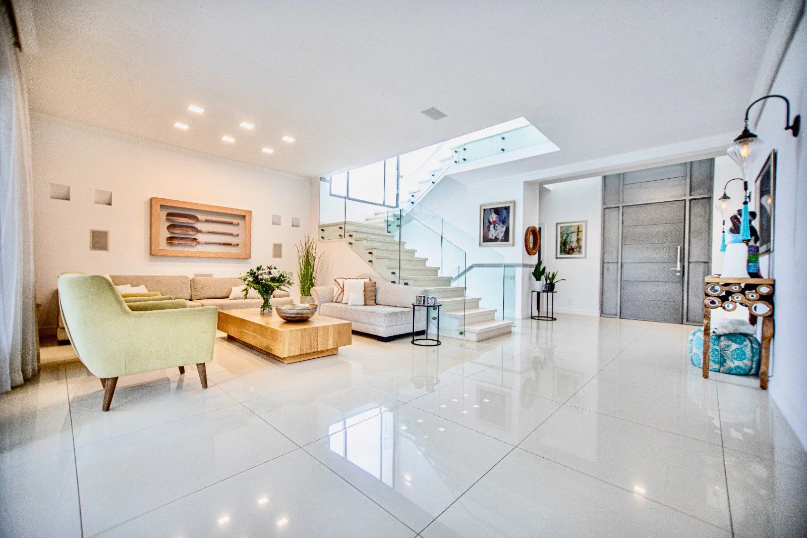 A modern, well-lit living room with white tiled floors, a staircase with glass railing, a beige sectional sofa, a light green armchair, a wooden coffee table with a flower vase, wall art, and decorative plants.