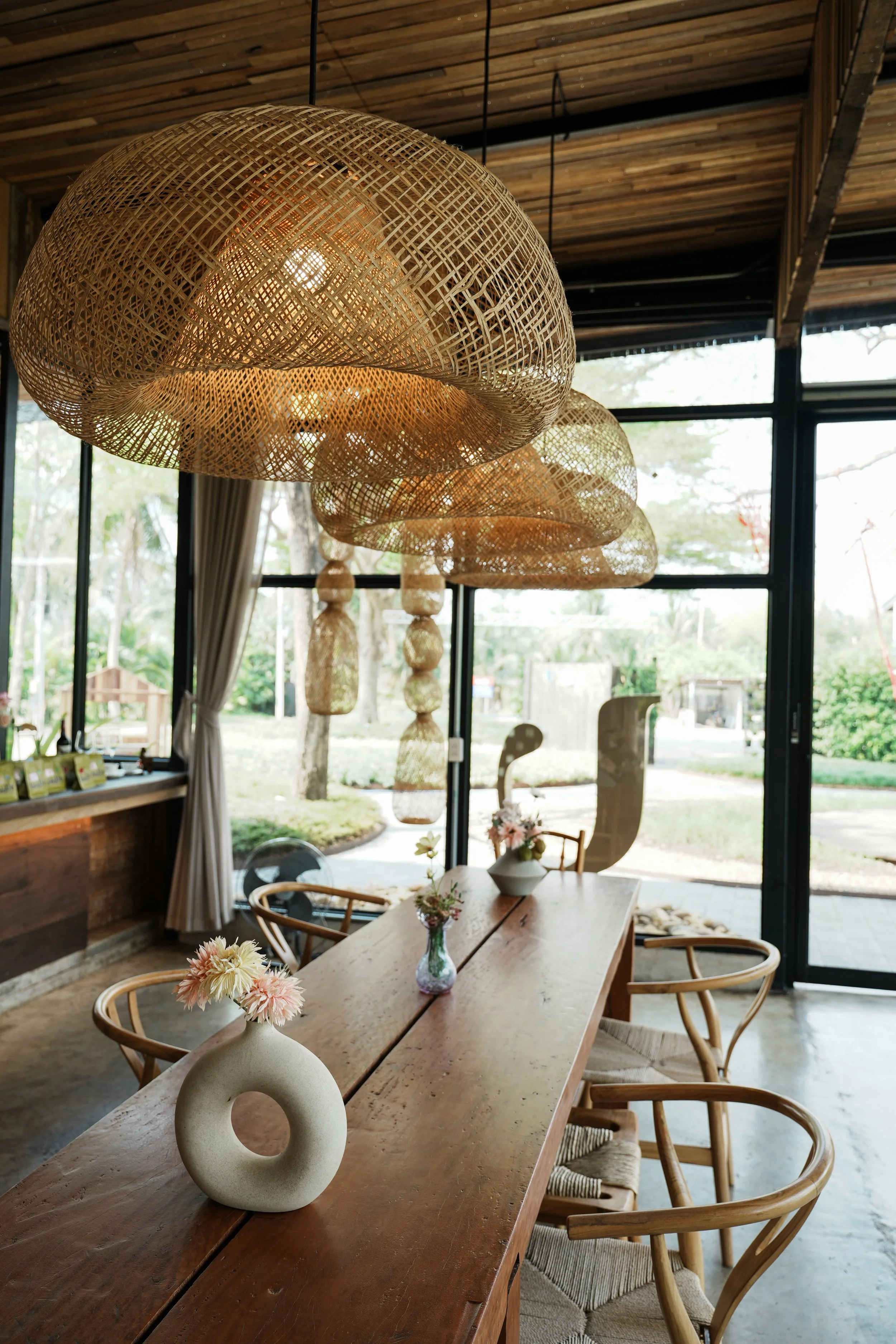 Interior of a modern dining area with wooden table and chairs, large woven pendant lights hanging from the ceiling, and large windows revealing greenery outside.