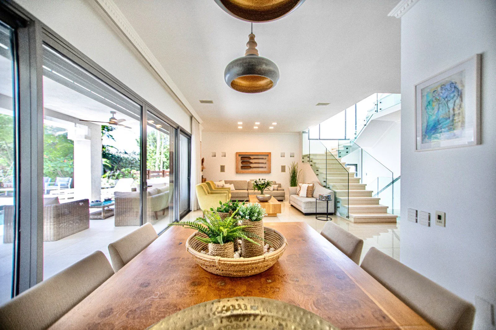 Bright modern living room with glass sliding doors leading to outdoor patio, featuring a dining table with potted plants, and stairs with glass railing leading upstairs.