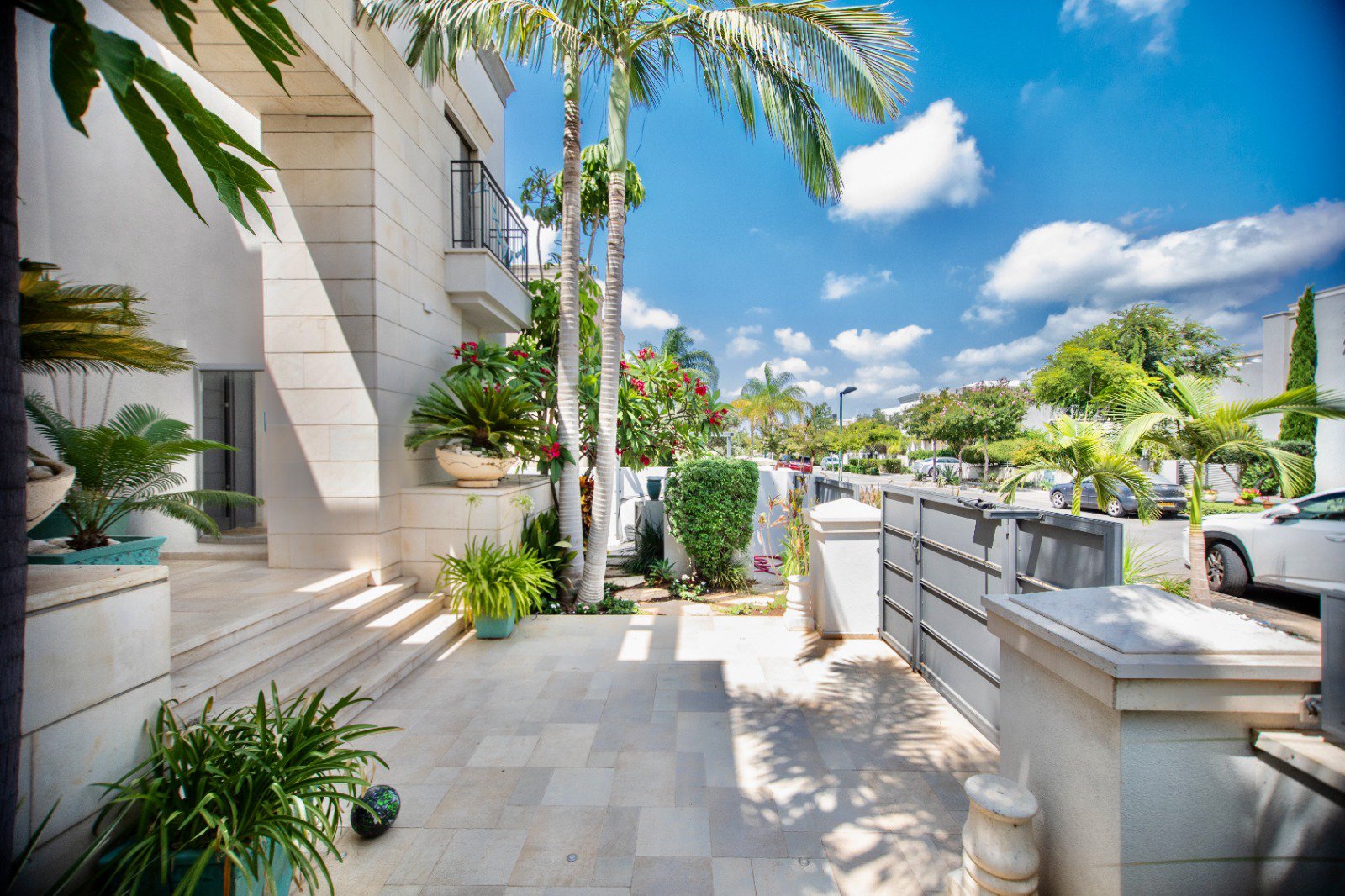Sunny outdoor patio with beige tiled flooring, potted plants, a cactus, palm trees, bushes, a modern white building with stairs, and a street view with parked cars under a blue sky with clouds.