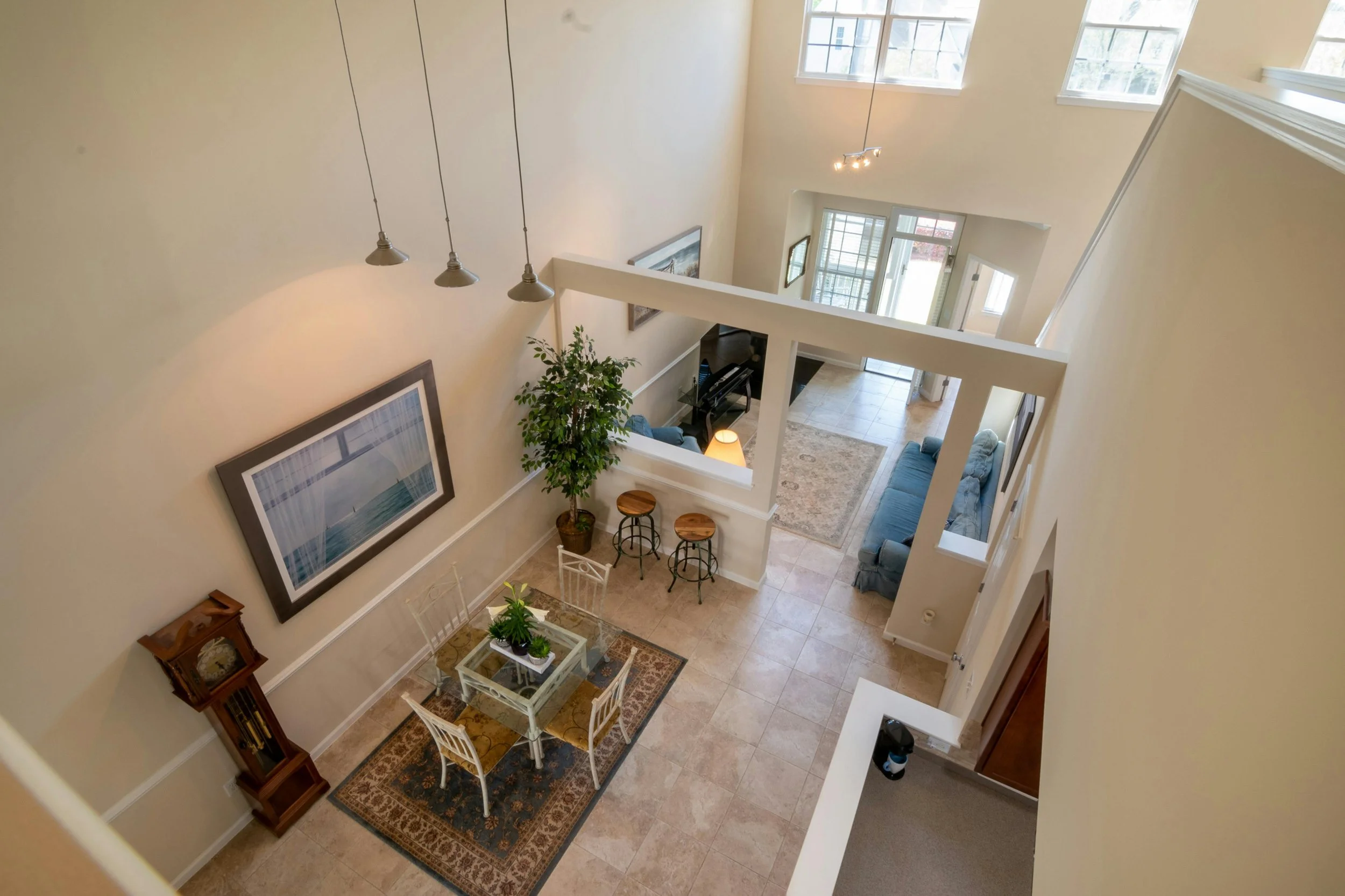 An aerial view of a spacious living room with a dining area, featuring a vase with green plants, a grandfather clock, a large framed artwork, a potted plant, a small bar table with two stools, a blue sofa, a piano, and a rug, with natural light comin