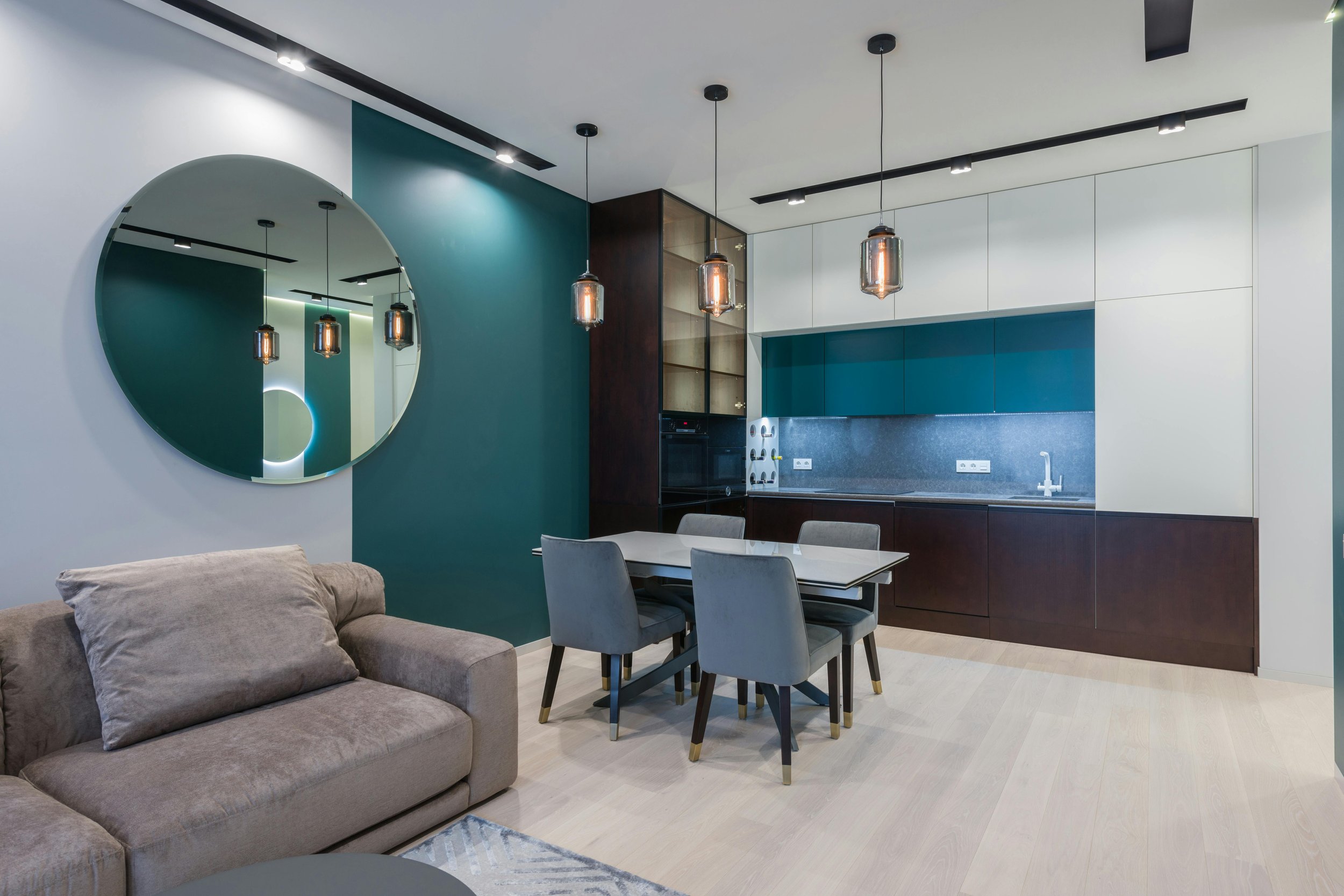 Open-concept living room and kitchen with beige sofa, dining table and four chairs, teal accent wall, modern pendant lights, and white and dark wood cabinetry.