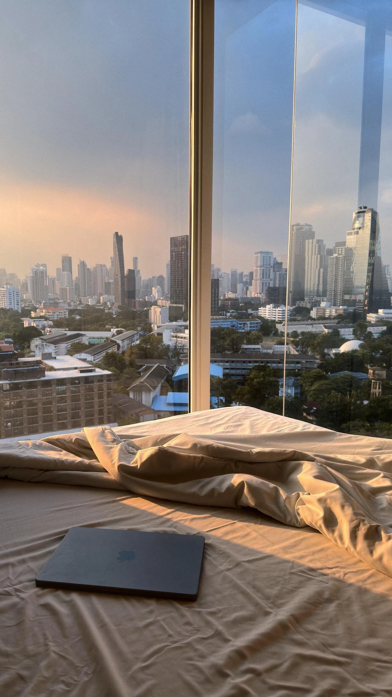 A laptop lying on a rumpled bedsheet in front of a large window showing a city skyline at sunset.