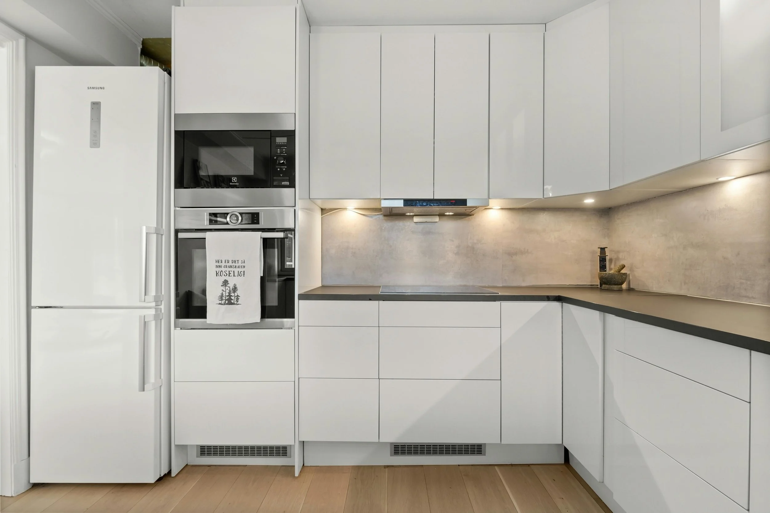 Modern white kitchen with built-in appliances, black countertops, and minimalist design. There's a refrigerator, microwave, and oven, with a towel hanging on the oven handle. The countertop has a small mortar and pestle and a soap dispenser. The kitc