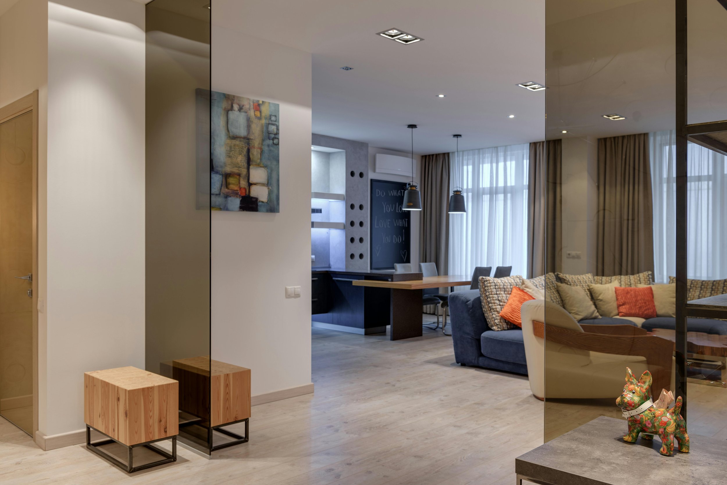 Modern living room with a blue sofa, patterned throw pillows, and a coffee table, separated by a glass partition, with dining area and kitchen in the background, natural light from large windows, and decorative accents including a colorful dog figuri