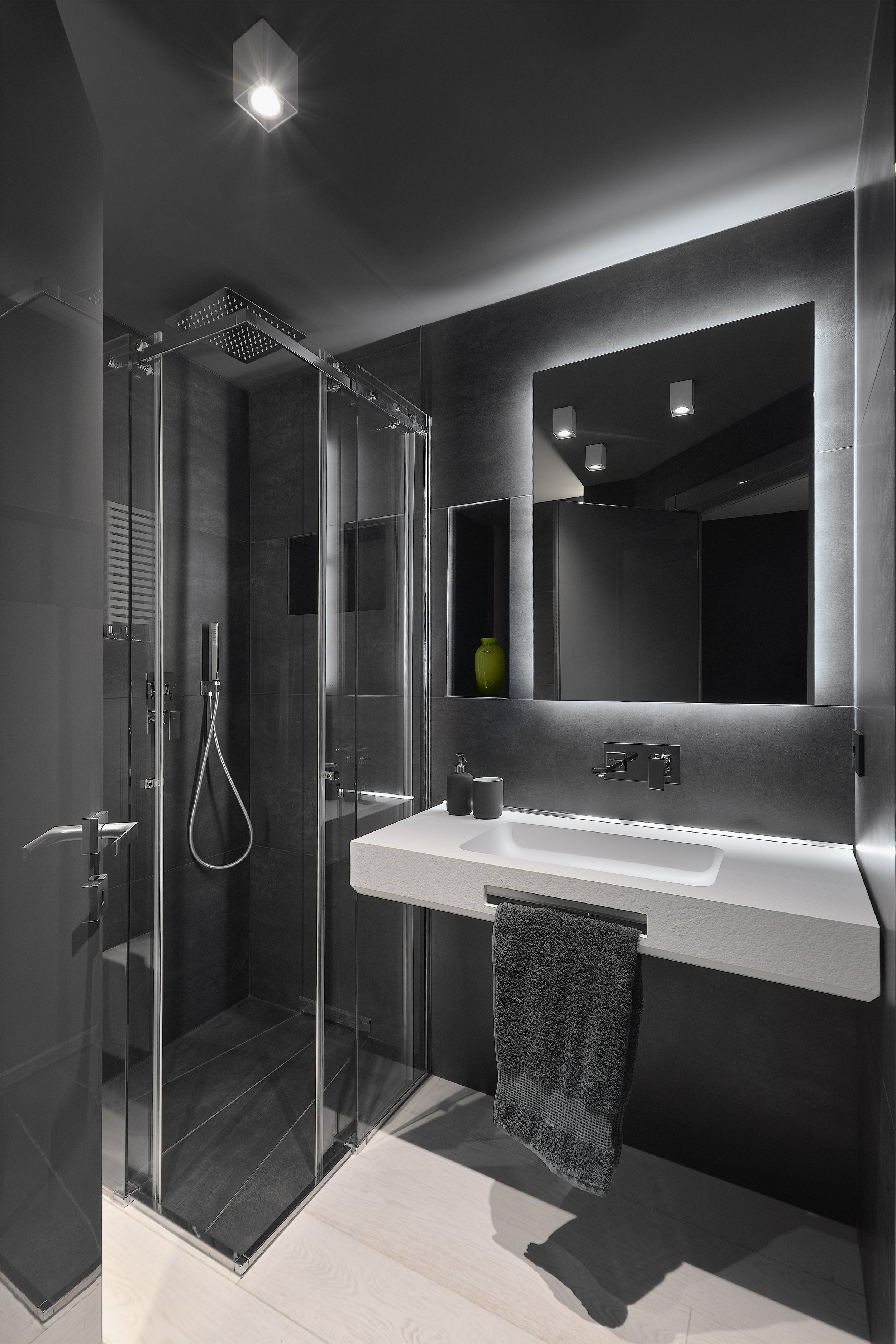 A modern, minimalist bathroom with dark tiled walls, a glass-enclosed shower with a rainfall showerhead, a white sink with a vanity, a large backlit mirror, and a black towel hanging on a rail.