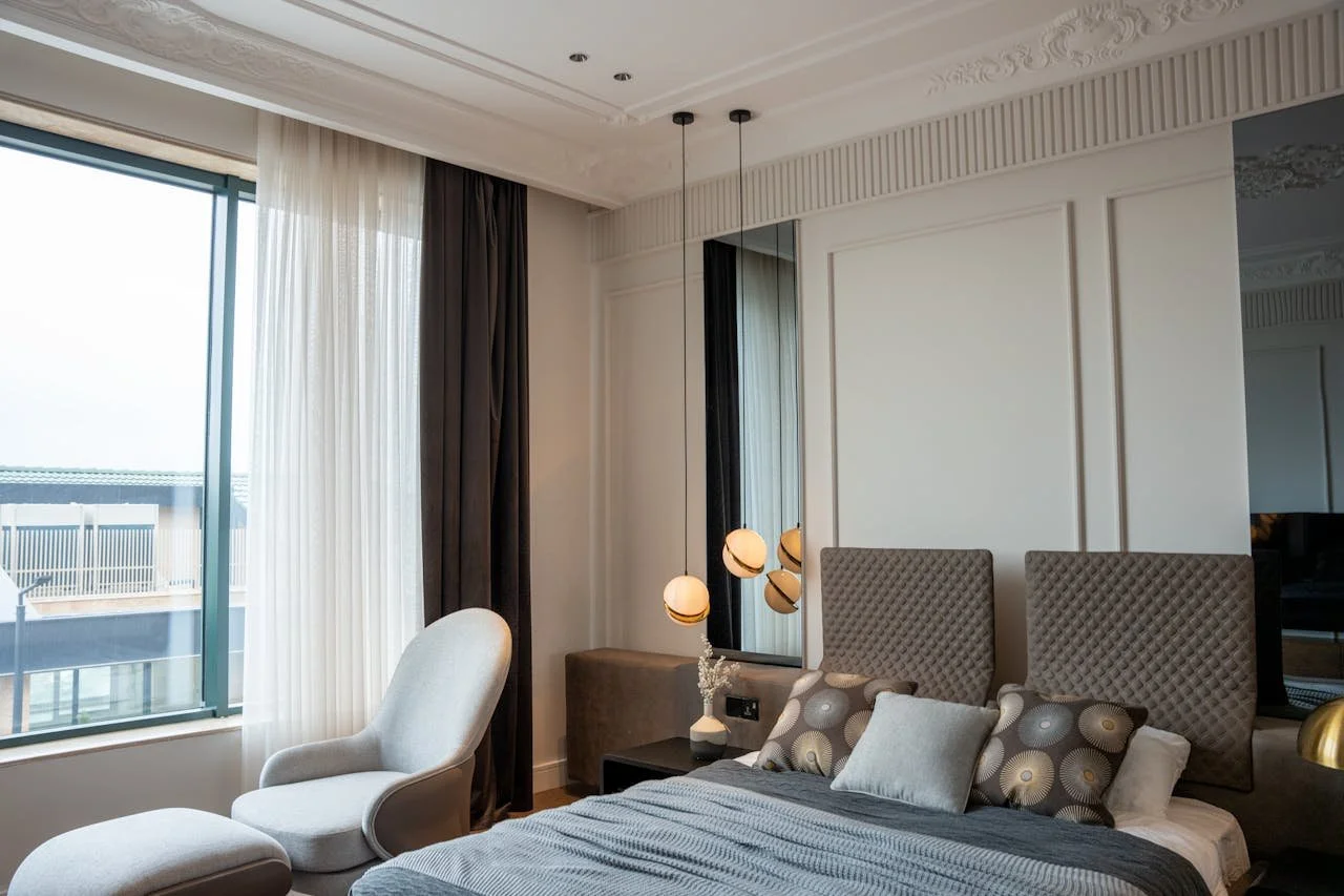 A modern hotel bedroom with a window view of neighboring buildings, a white armchair with an ottoman, a bed with decorative pillows, and stylish hanging pendant lamps.
