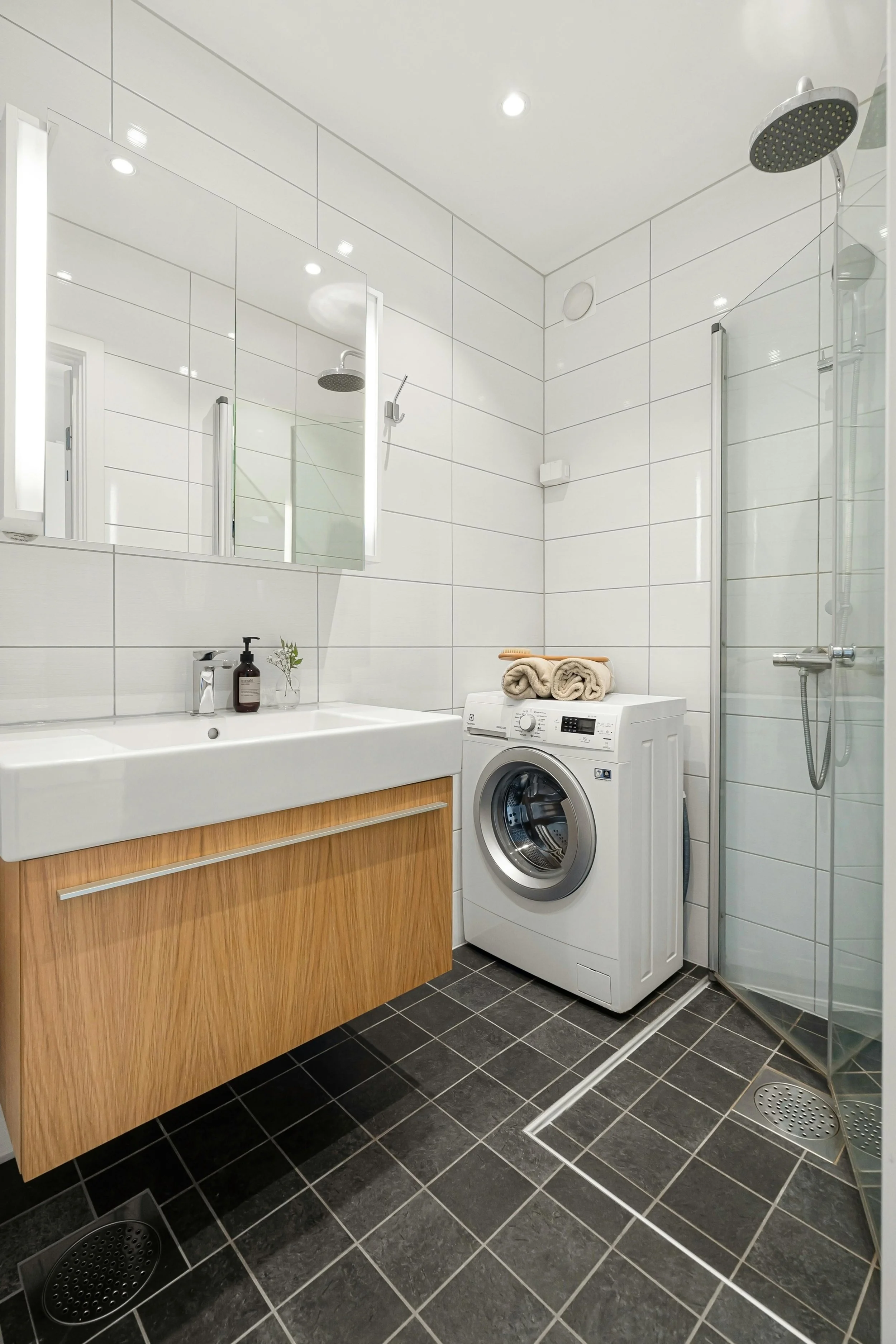 Modern bathroom with white tiled walls, black tiled flooring, a wooden vanity with a white sink, a mirror with lighting, a washing machine with folded towels on top, and a glass-enclosed shower.