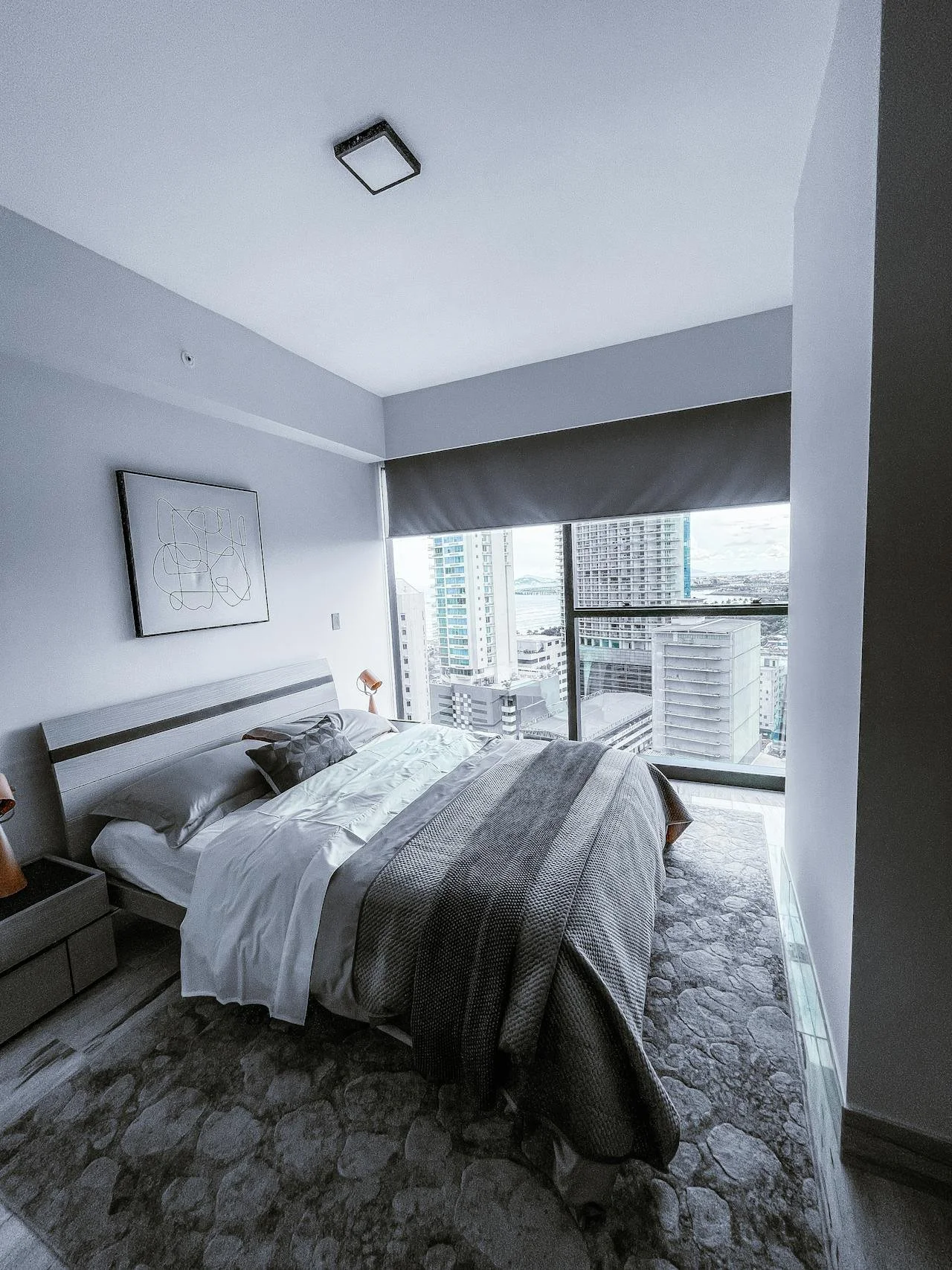 Modern bedroom with a large bed, gray bedding, and an abstract wall art piece, high-rise city view through large windows.