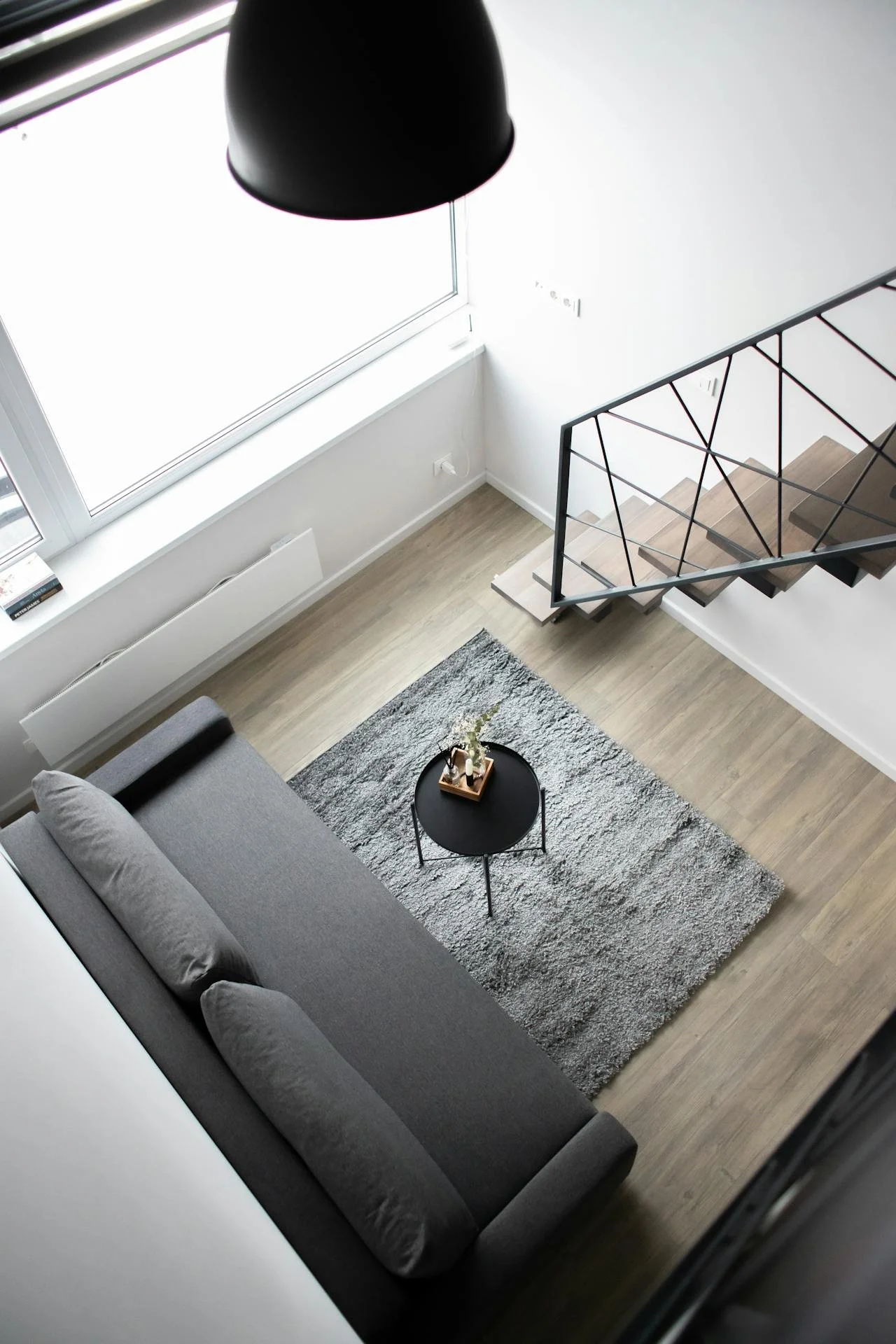 A minimalist living room viewed from above, featuring a gray sofa, a small black round coffee table with decorative items, a gray textured area rug, a large window with a white windowsill, and a staircase with wooden steps and a black metal railing.