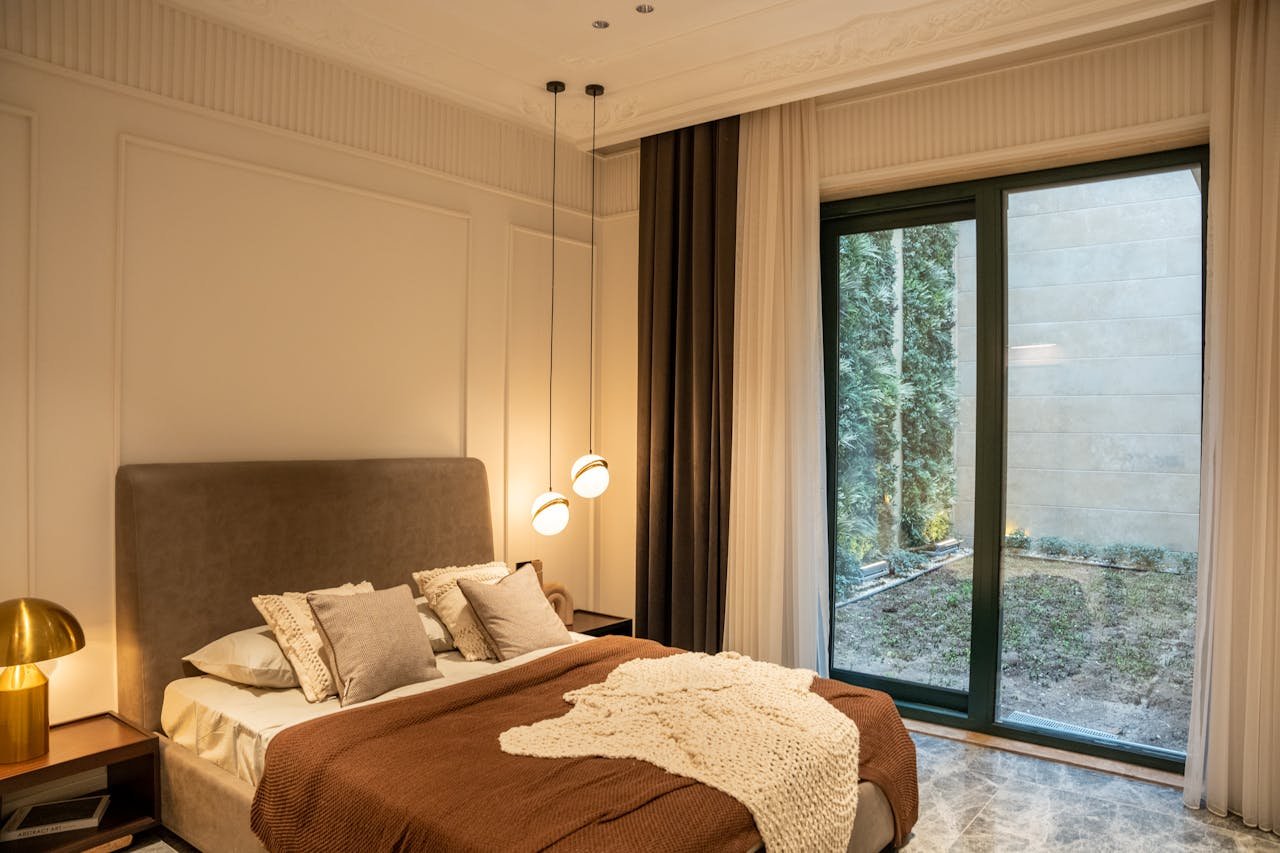 A bedroom with a beige upholstered bed, gray and beige pillows, brown blanket, and a cream knitted throw. There is a dark wooden nightstand with a gold lamp, beige curtains, a large glass sliding door showing trees outside, and tile flooring.