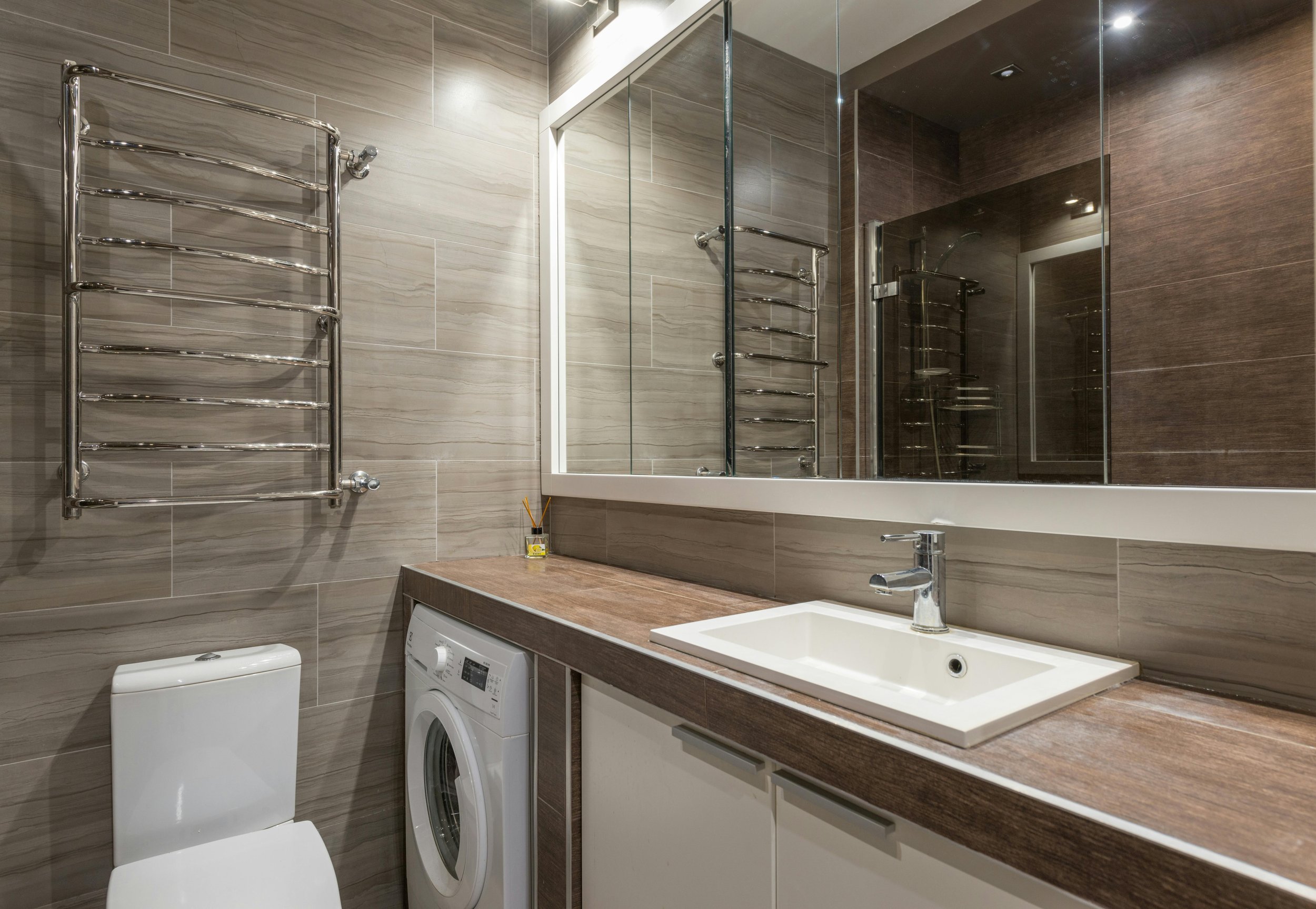 Modern bathroom with a large mirror, sink, washing machine, toilet, and heated towel rack, featuring beige and brown tiled walls.