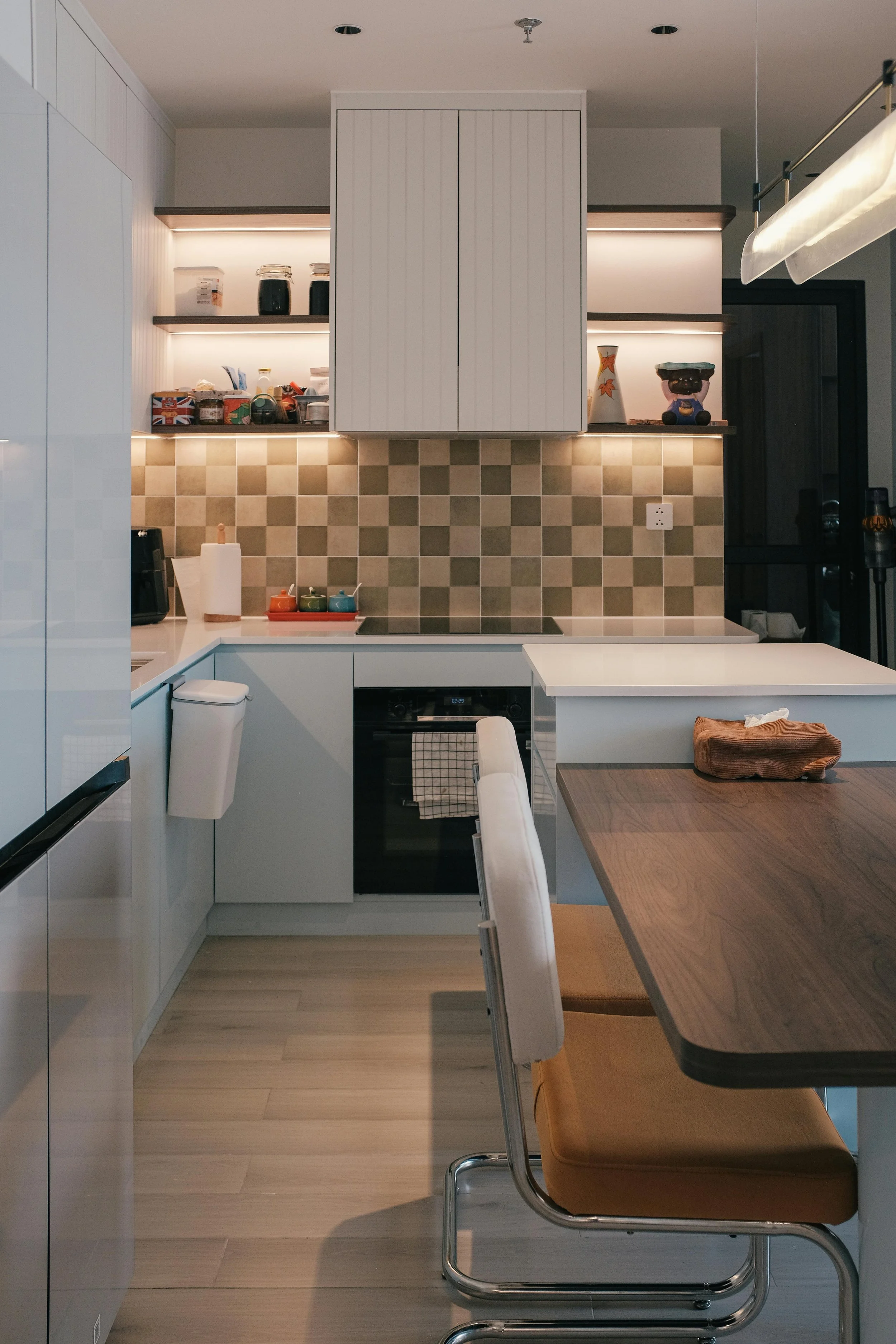 Modern kitchen with white cabinetry, beige checkered backsplash, open shelves with jars and decor, and a wooden table with beige and brown chairs.