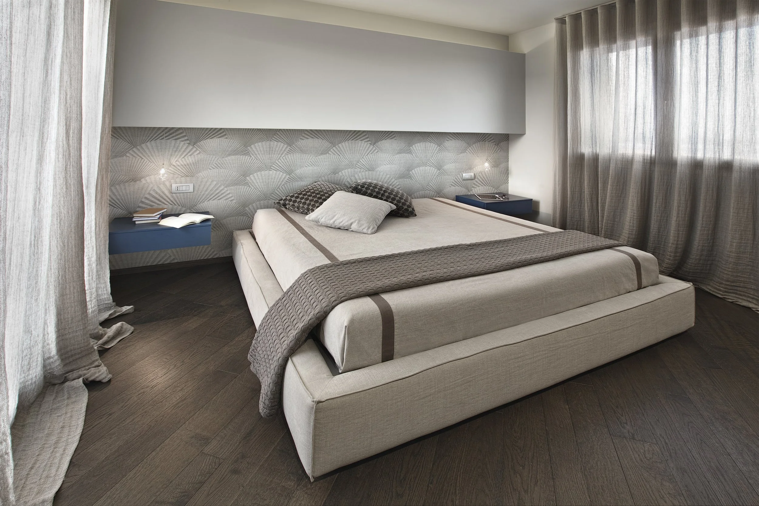 Modern bedroom with a large bed, two nightstands with books and a tray, and patterned wall behind the headboard. The room has beige and brown tones with sheer curtains covering the windows.