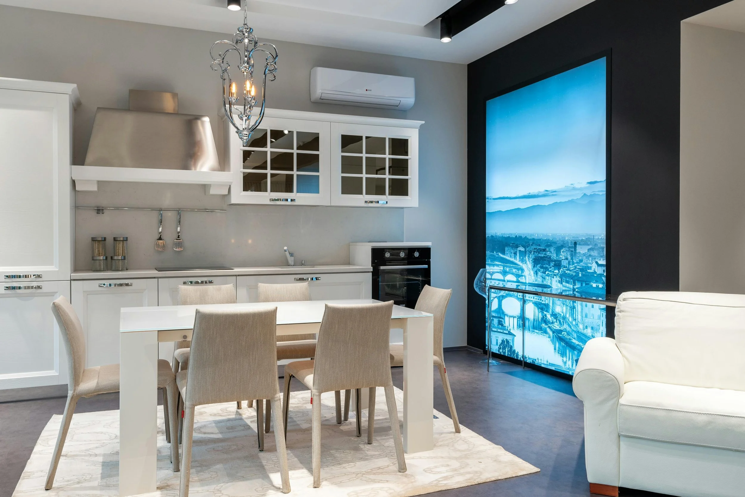 Modern kitchen and dining area with white cabinets, a dining table with six beige chairs, a chandelier, a large digital screen showing cityscape, and a white sofa.