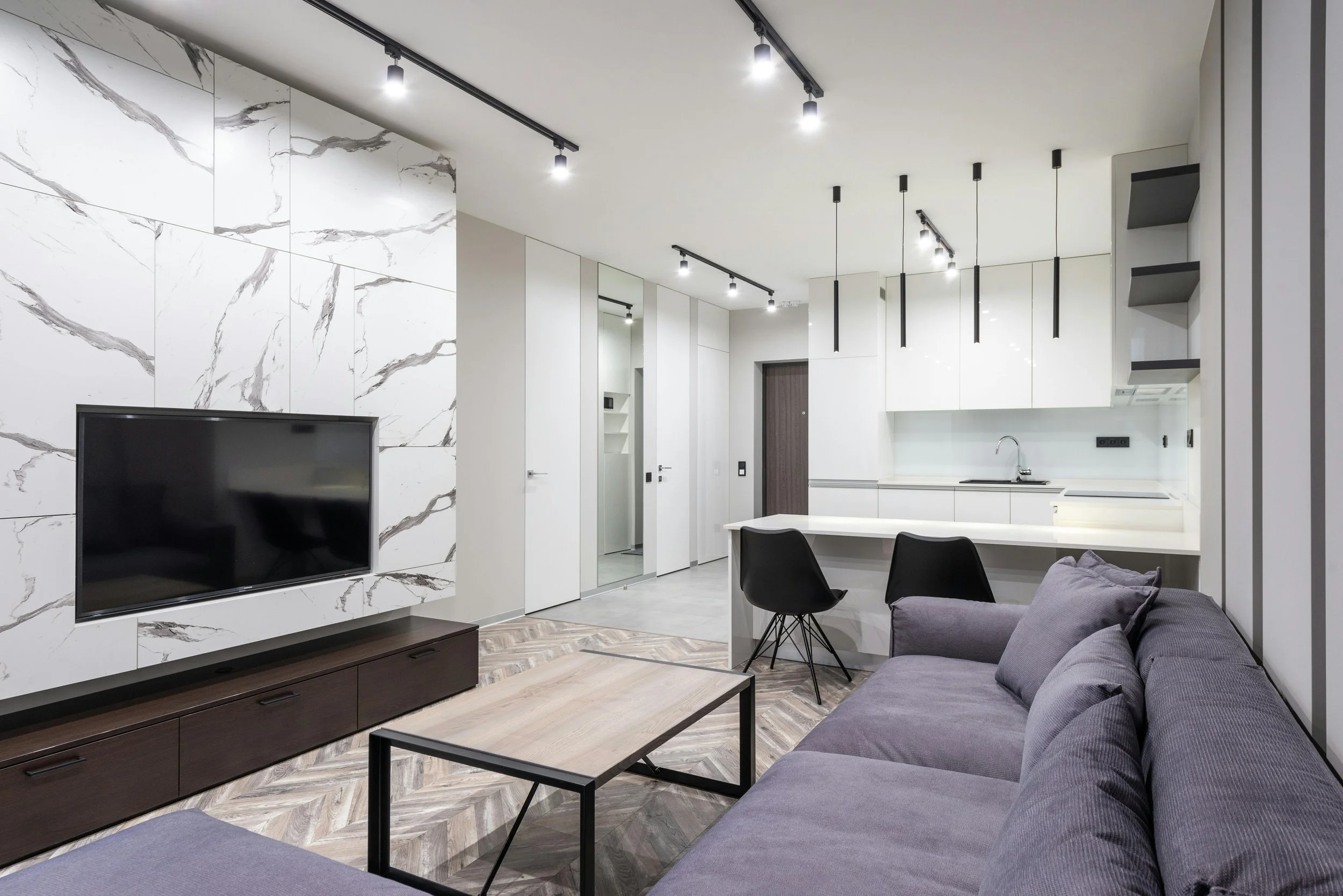Modern apartment living room with a purple sofa, wooden coffee table, TV on a marble wall, and an open kitchen with white cabinets and black pendant lights.