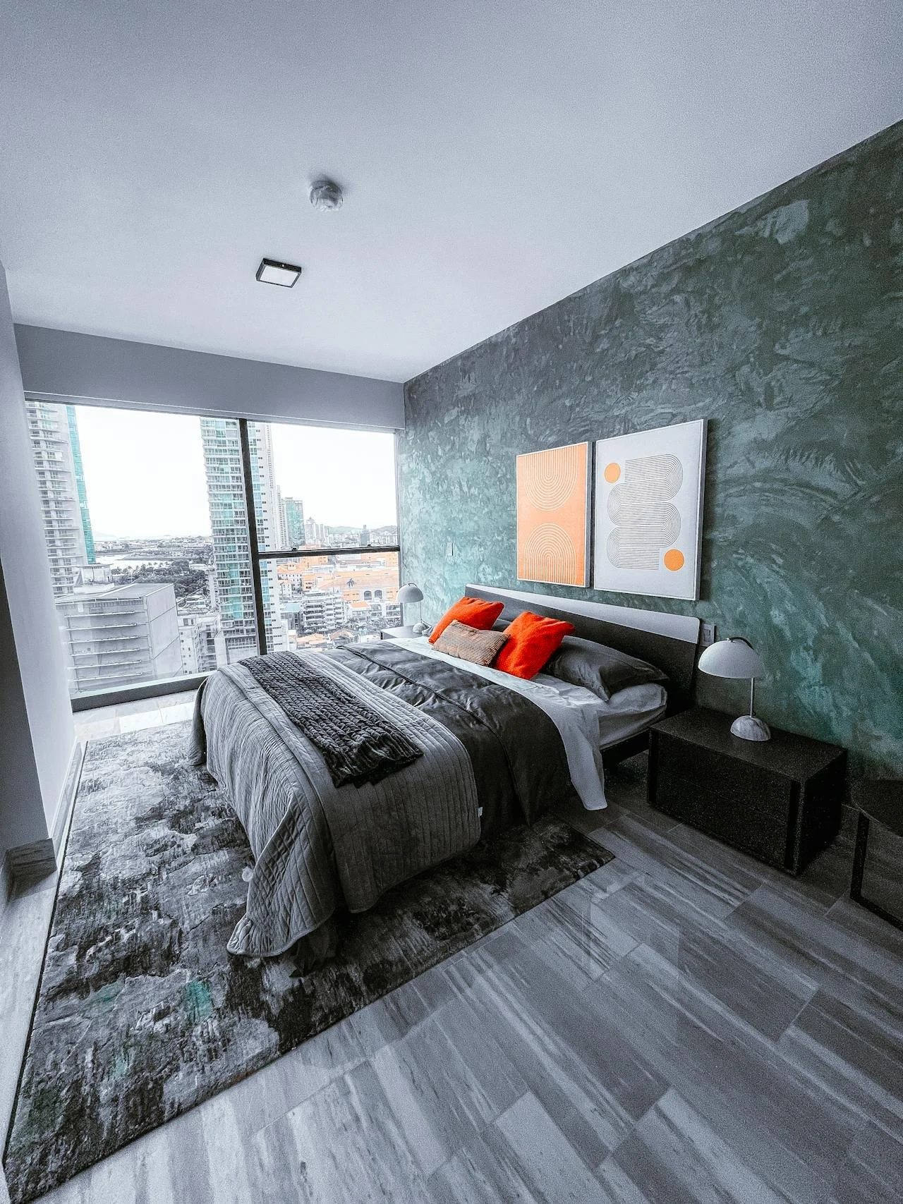 Modern bedroom with large window showing city skyline, bed with gray bedding and orange accent pillows, abstract art on green textured wall, and dark nightstands.