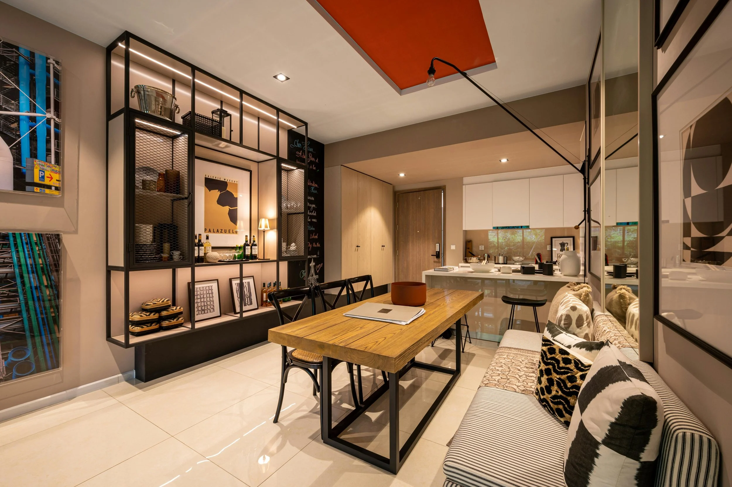 Modern open-plan dining and kitchen area with a wooden table, black chairs, beige and patterned cushions on a striped and animal print sofa, and decorative shelving and artwork on the wall.