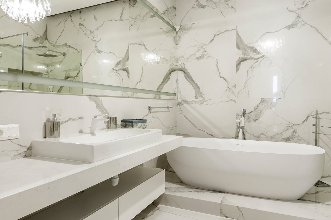 Modern bathroom with white marble walls and flooring, a white bathtub, a white rectangular sink on a countertop, wall-mounted faucet, mirror, and minimalist decor.