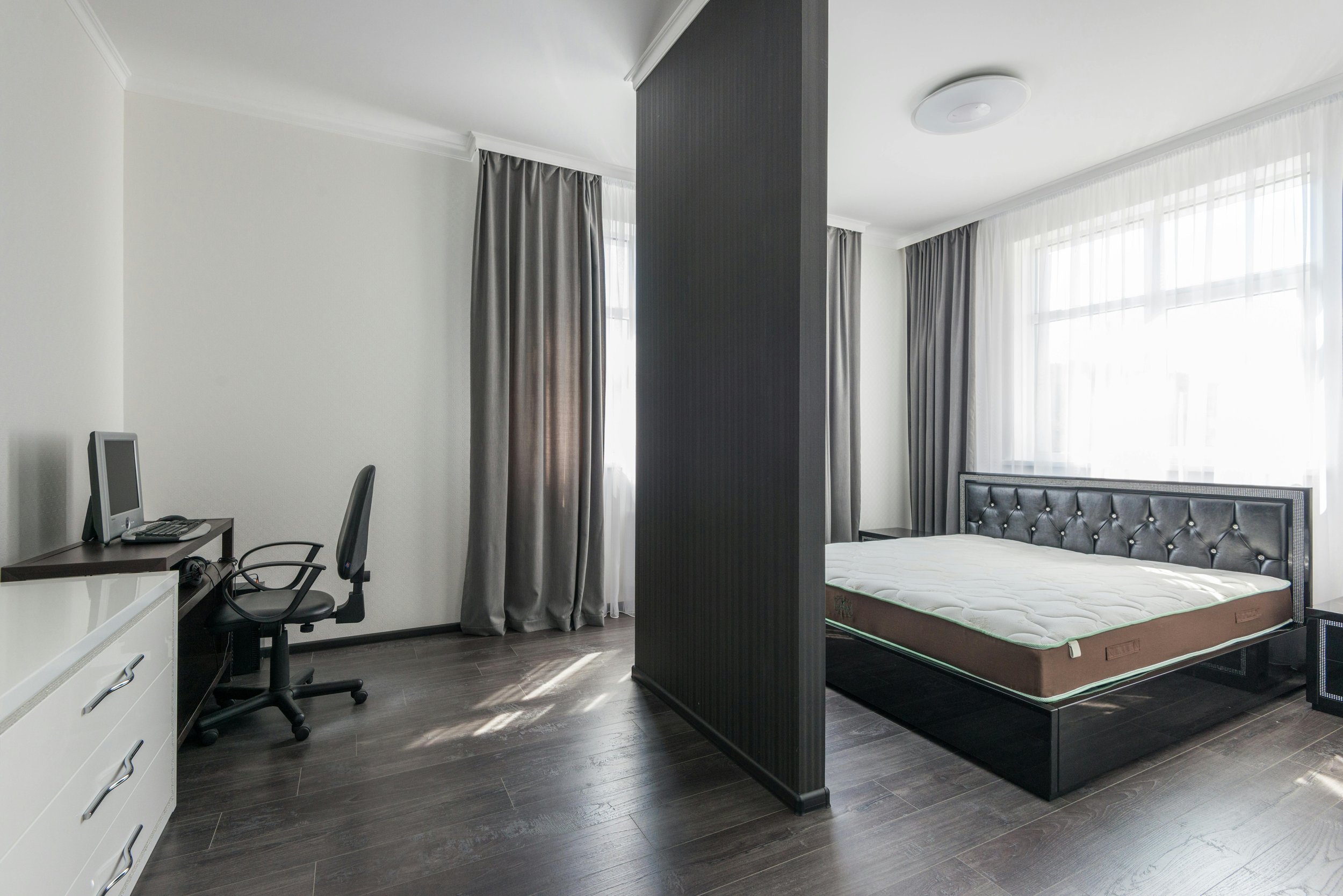 Spacious modern bedroom divided by a black partition wall, with a bed on the right side, large windows with curtains, and a small desk with a computer and chair on the left side.