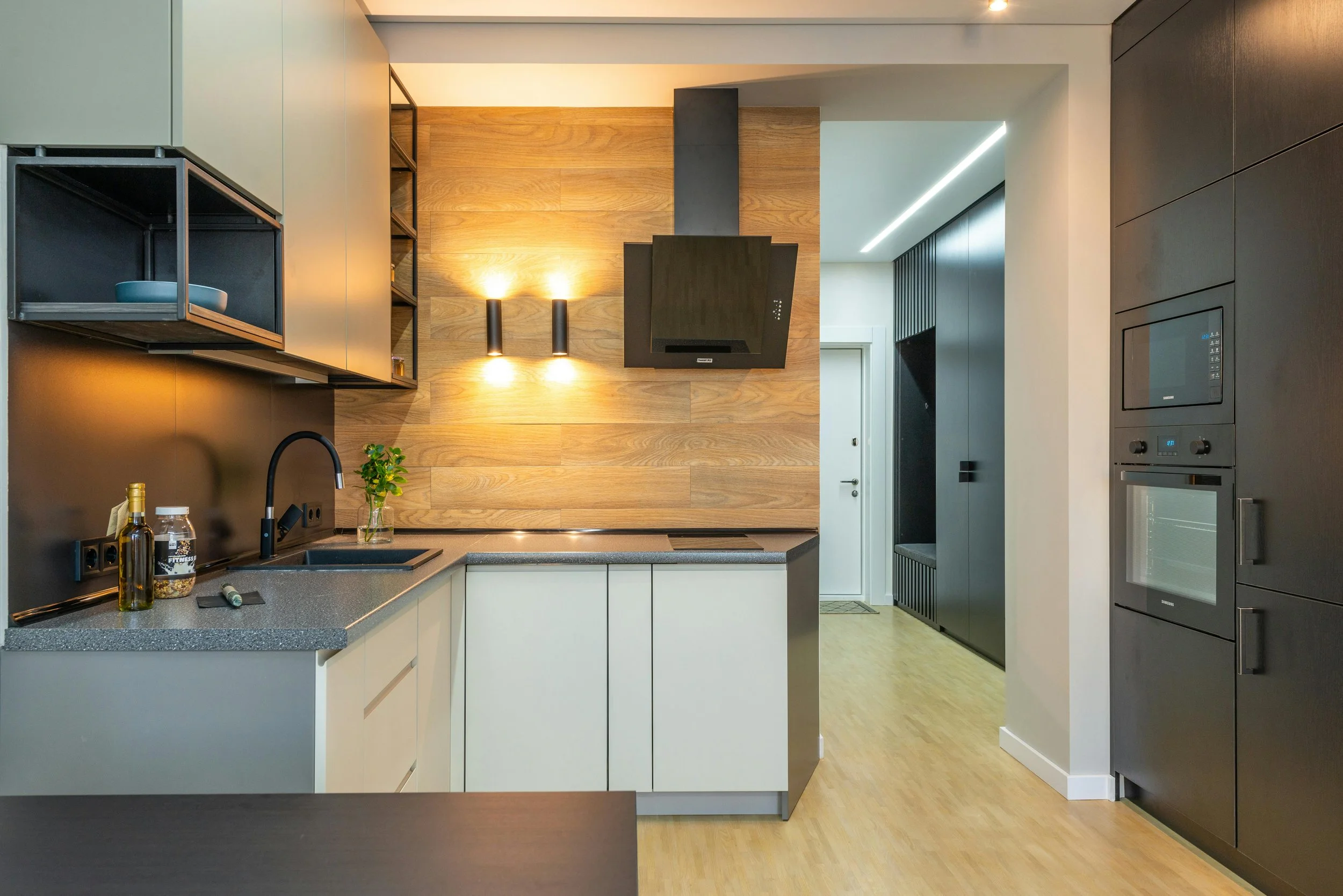 Modern kitchen with black and white cabinets, gray countertops, a black sink with a plant nearby, a black range hood, and a wooden accent wall with wall-mounted lights. There is a microwave and oven built into the black cabinetry on the right.