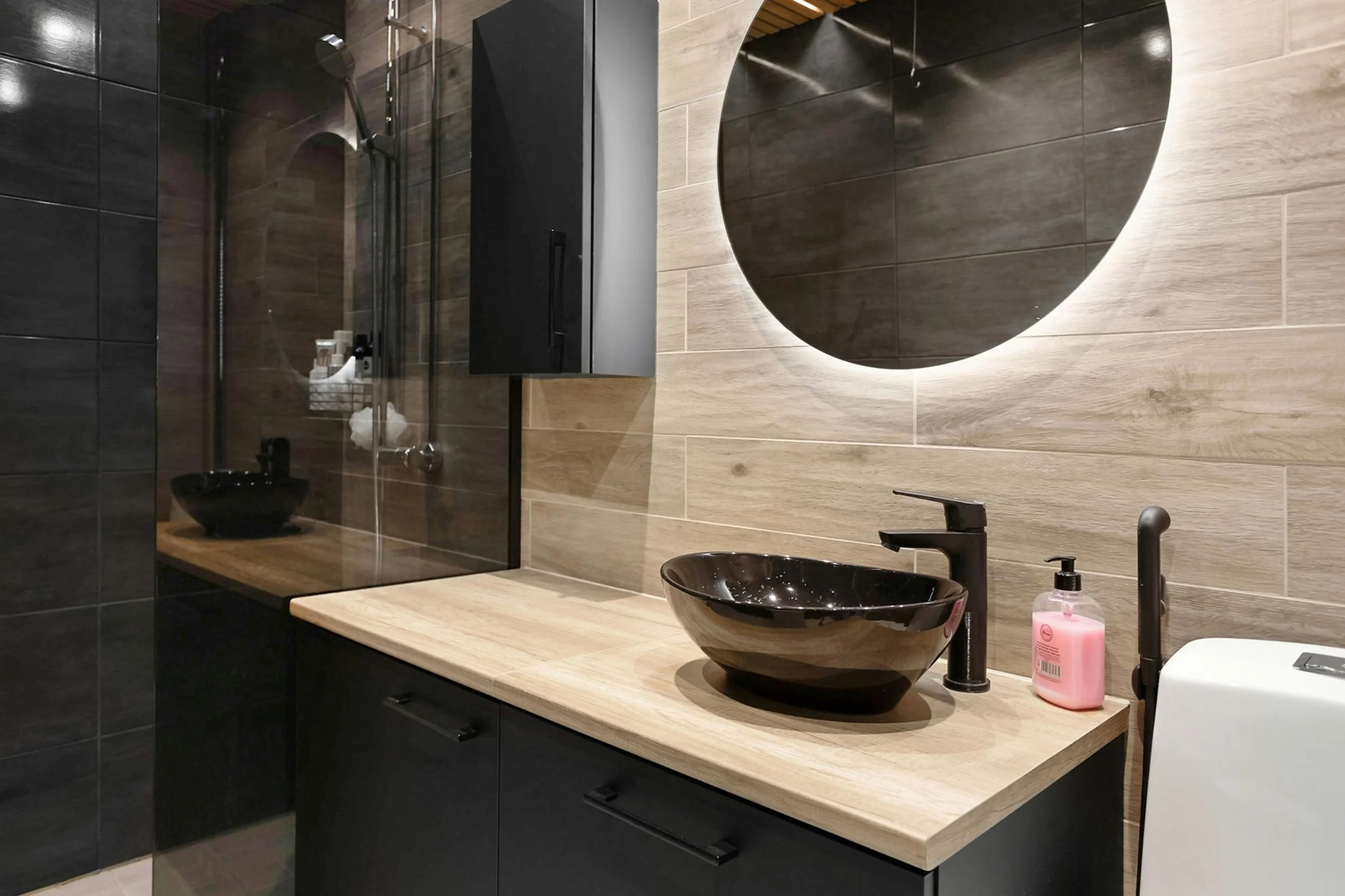 Modern bathroom with black vessel sink, black faucet, pink liquid soap, round mirror with backlighting, beige wood countertop, dark lower cabinets, and a partially visible shower with black fixtures.