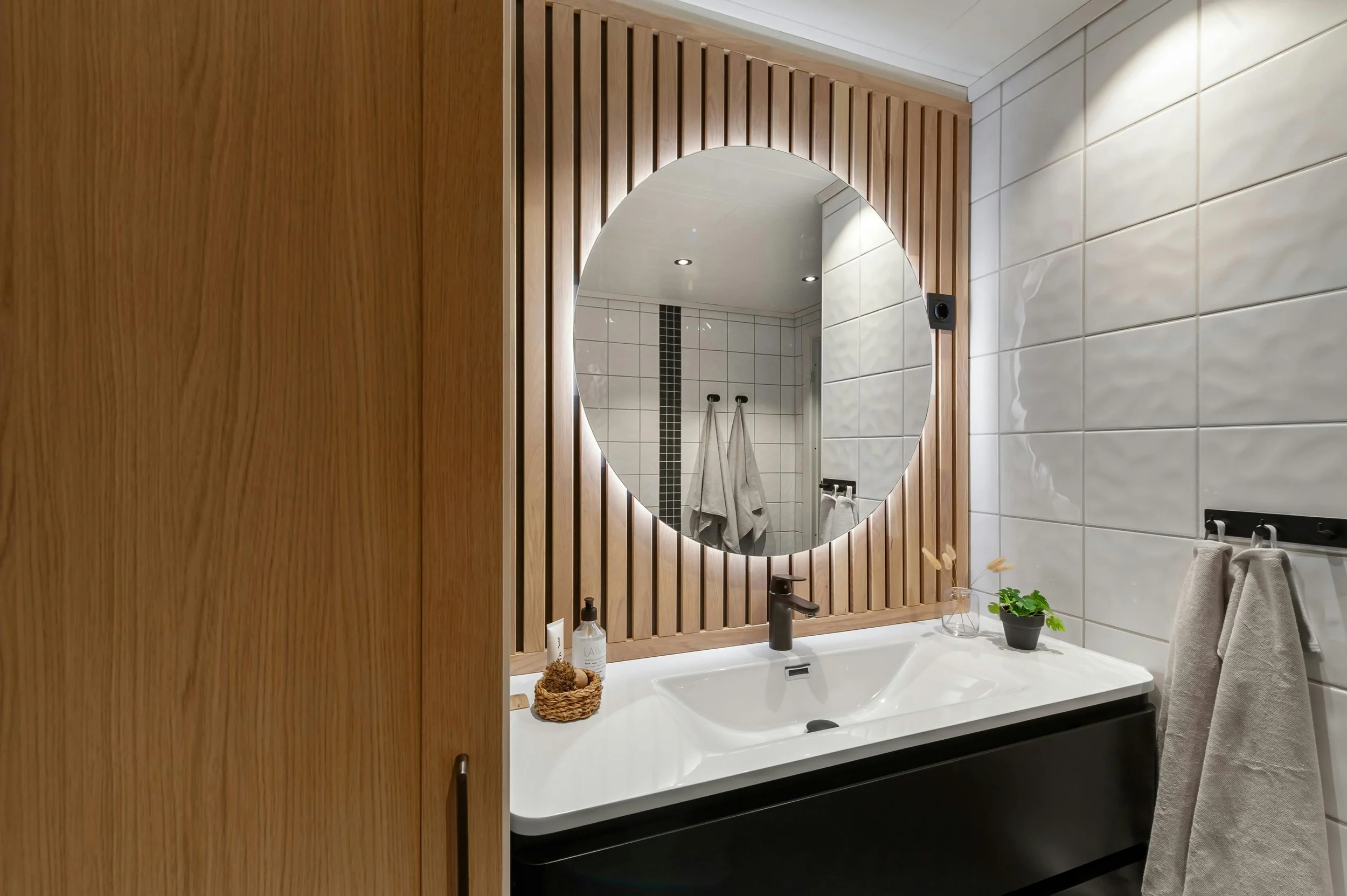 Modern bathroom with a large round mirror above a white sink, wooden panel wall behind the mirror, and tiled wall to the right with two towels hanging.