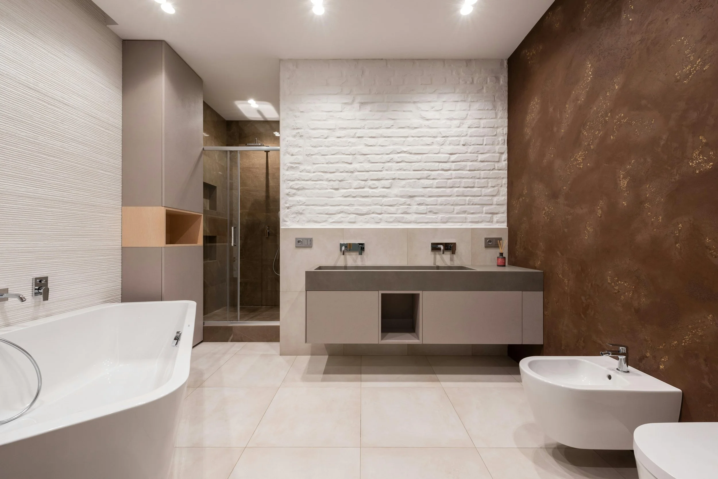 Modern bathroom with a white bathtub, a walk-in shower with brown tiles, a gray vanity with dual sinks, a white brick accent wall, and a textured brown painted wall.