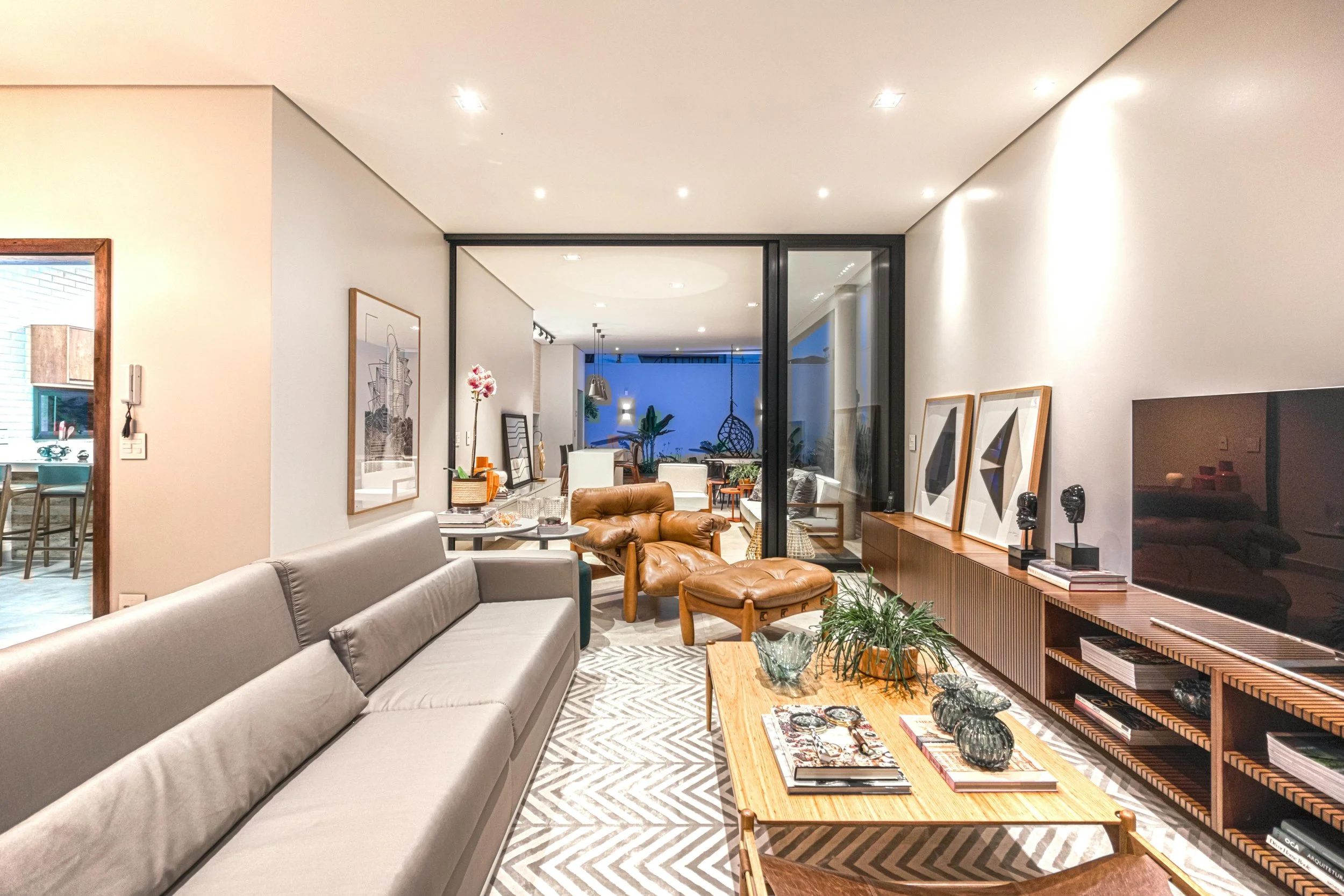 Modern living room with a gray sofa, wooden coffee table, leather armchair, and decorative sculptures and artwork on a wooden console, with a large flat-screen TV and glass sliding doors leading to a patio.