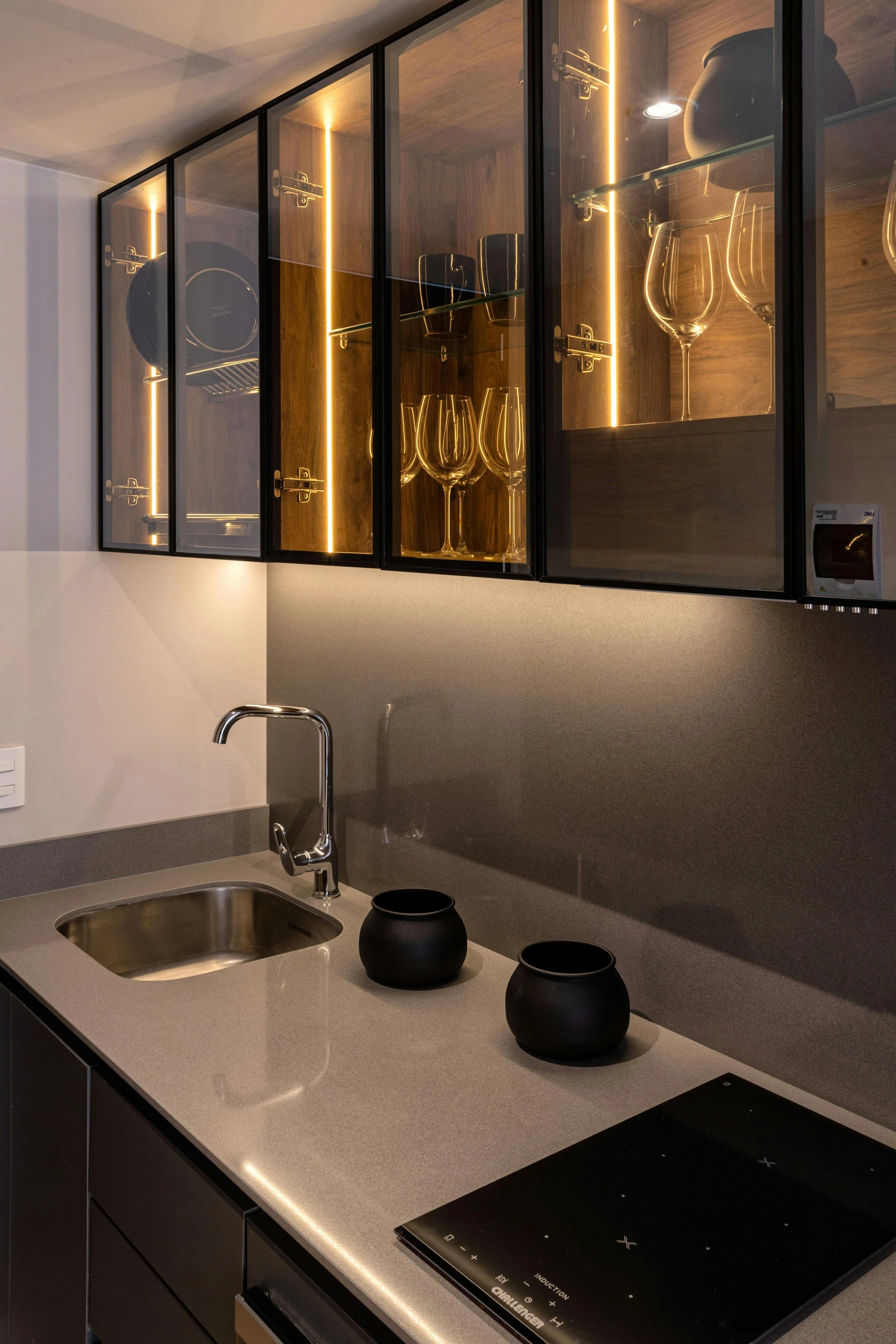 Modern kitchen with upper glass cabinet displaying wine glasses and dark pottery, a gray countertop with black vases, a faucet, a sink, and an electric stove.