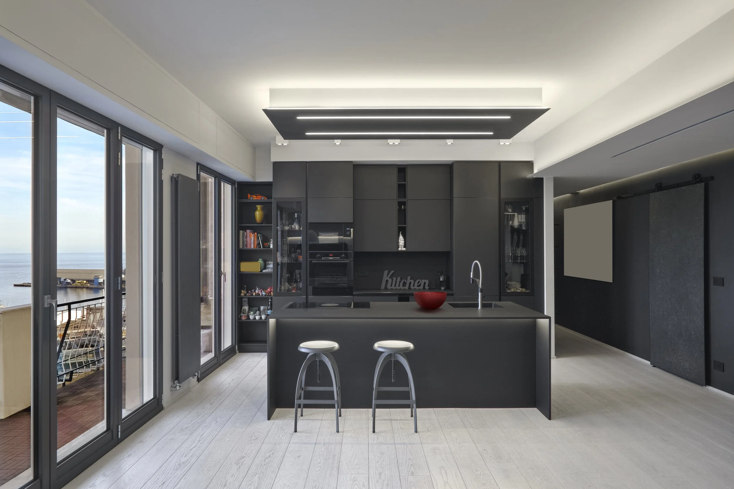 Modern kitchen with black cabinetry, an island with two stools, large windows with a view of the water, and a ceiling with sleek lighting fixtures.