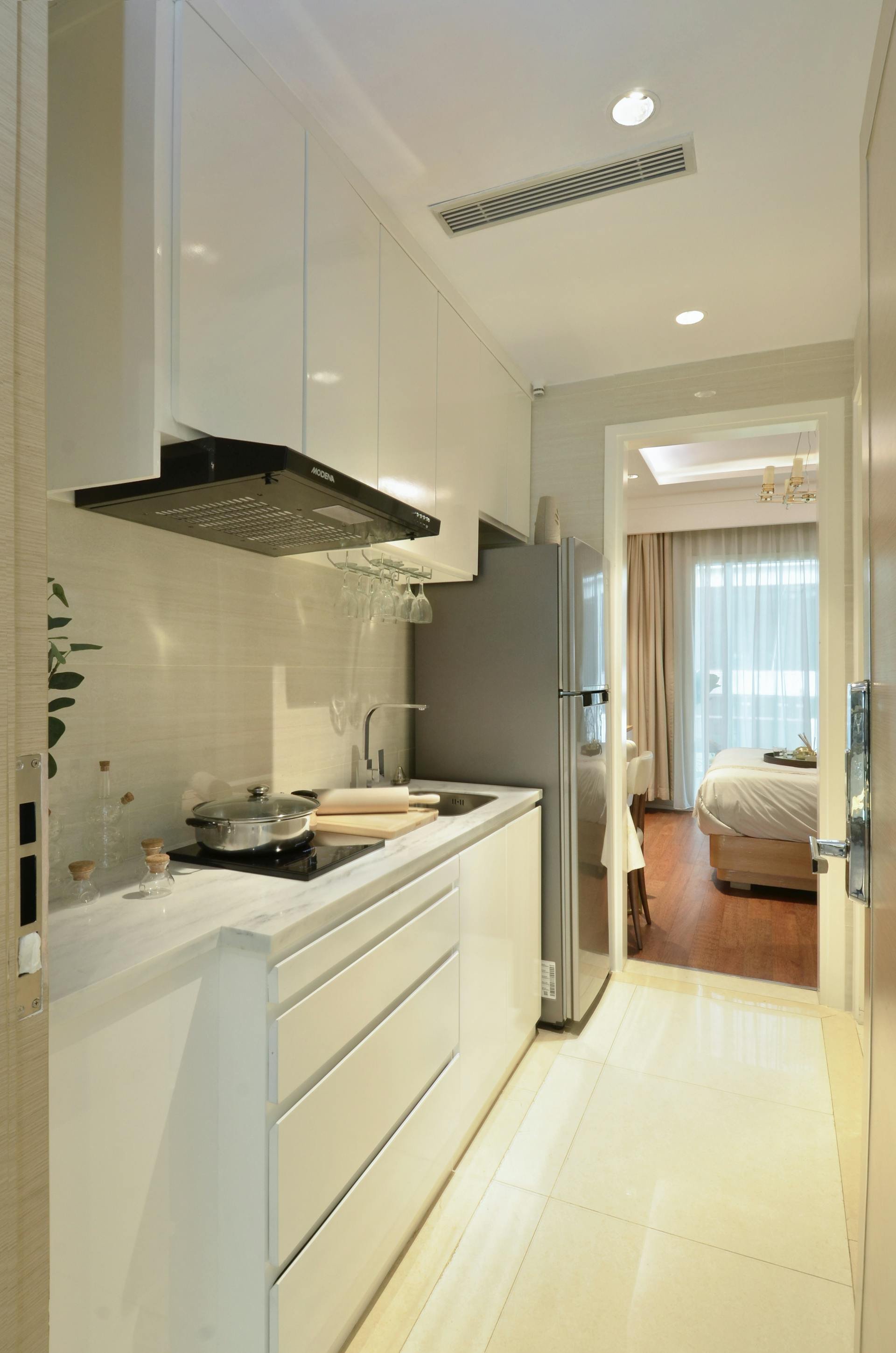 Modern kitchen with white cabinets, a countertop with a cooking pot, glass cups hanging, and a refrigerator, with a view into a bedroom with a bed and large window.