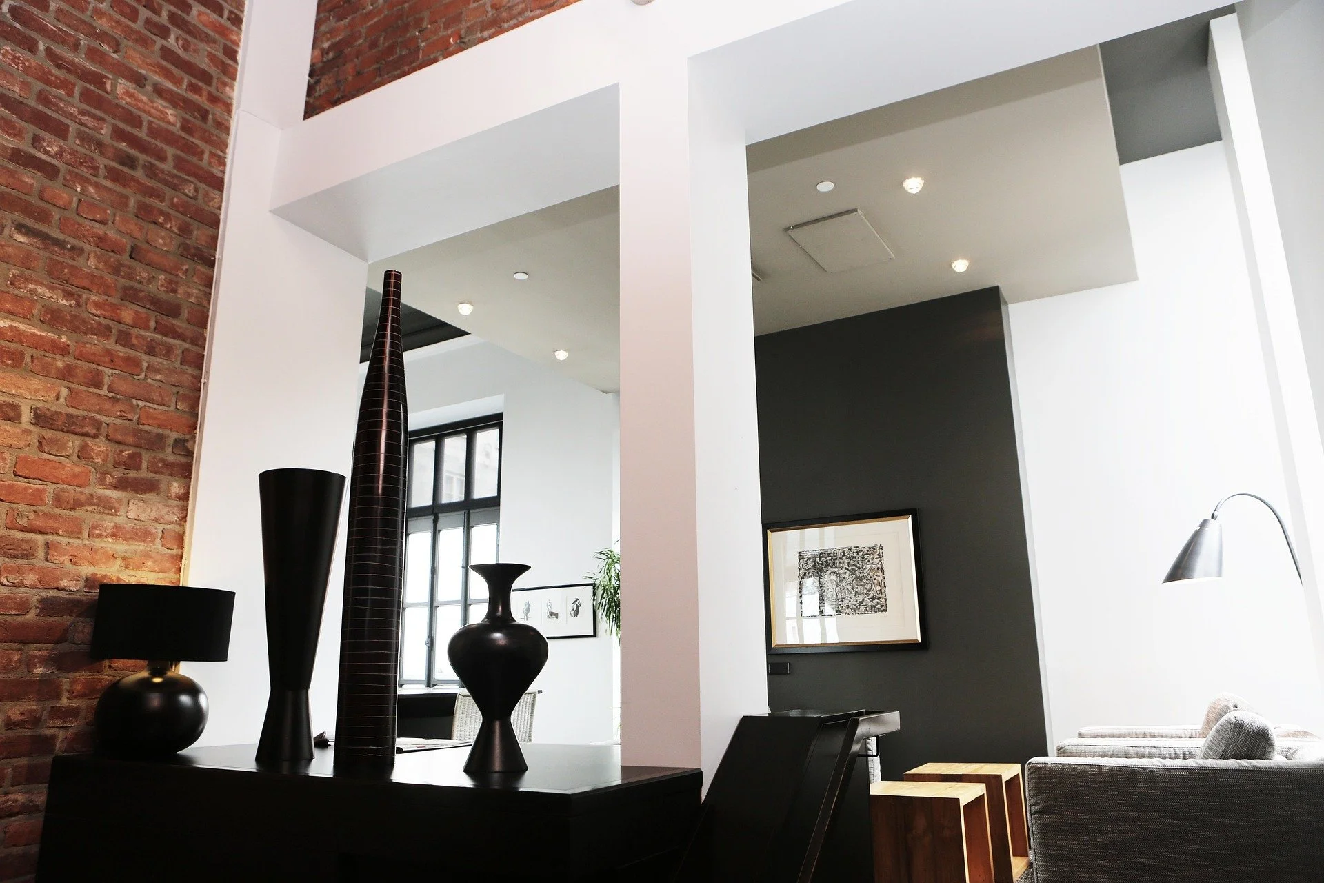 Contemporary living room with exposed brick wall, black and white furniture, large windows, framed artwork, and decorative vases.