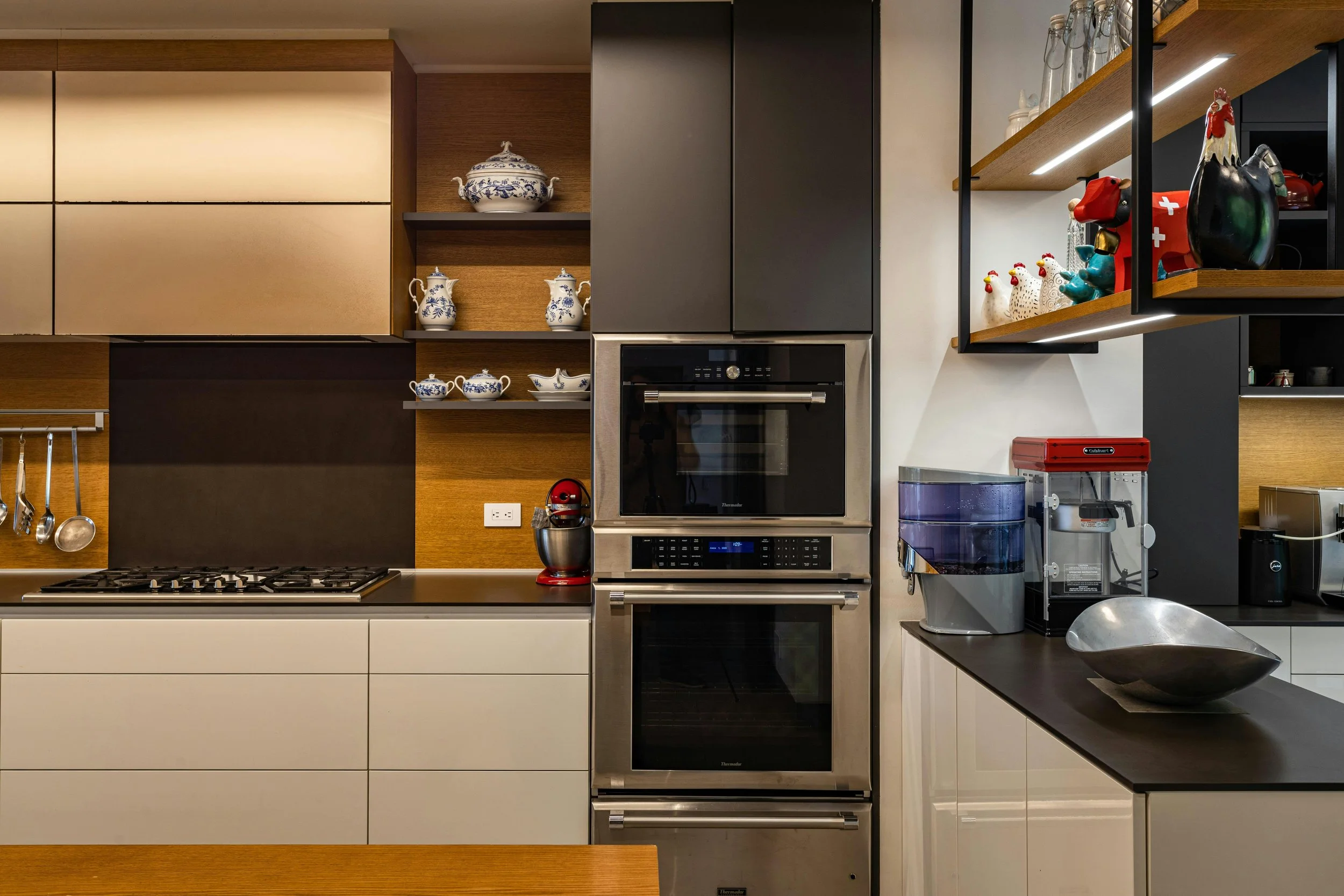 Modern kitchen with built-in double oven, wooden shelves with decorative dishes, black and white cabinetry, and kitchen appliances such as a coffee machine and water filter, with decorative rooster and cow figurines on open shelves.