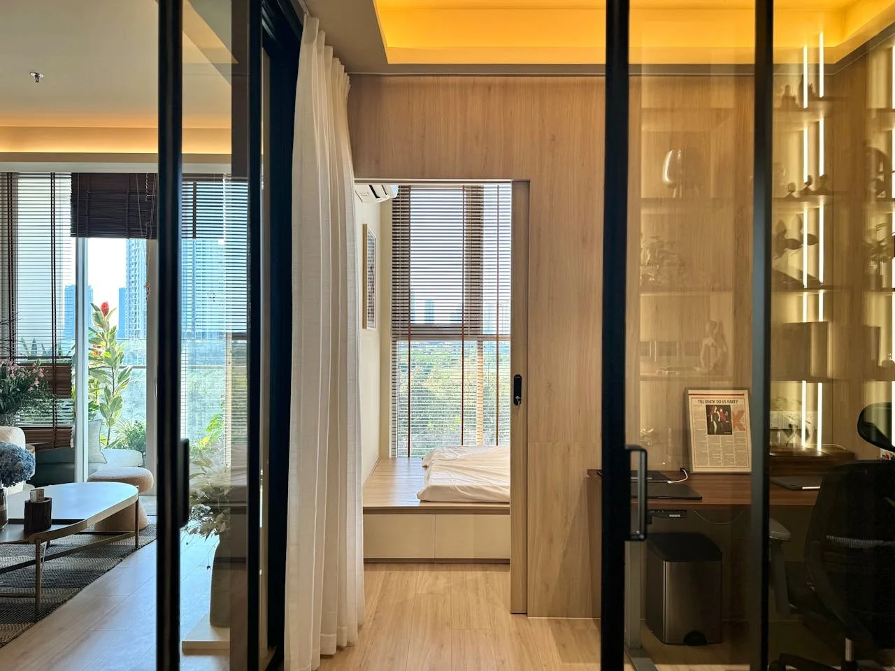 Interior view of a modern apartment or hotel room with a separated sleeping area, large windows with blinds, a small workspace with a desk, and a cozy living space with seating and plants.