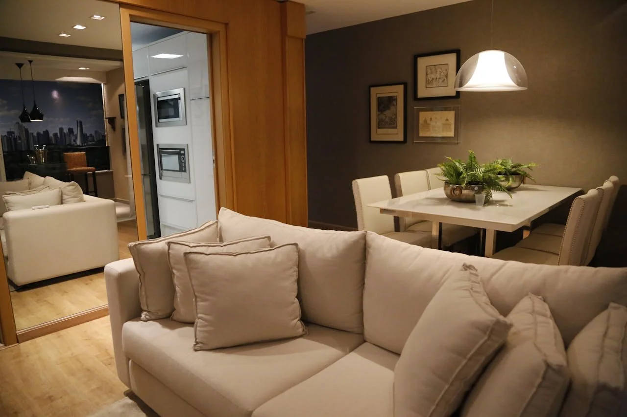 Living room with a white sofa and living room with a dining area with a white table and beige chairs, wall art, and a hanging light. Open doorway reveals a kitchen with appliances.