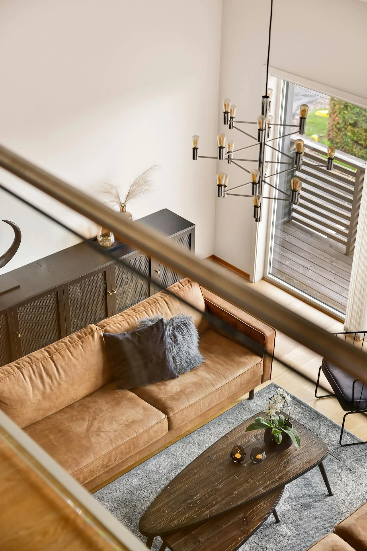 Top-down view of a modern living room with a tan sofa, a dark wooden coffee table, a gray area rug, black chair, and large glass sliding door leading to an outdoor deck. A black chandelier with exposed bulbs hangs from the ceiling, and a black cabine