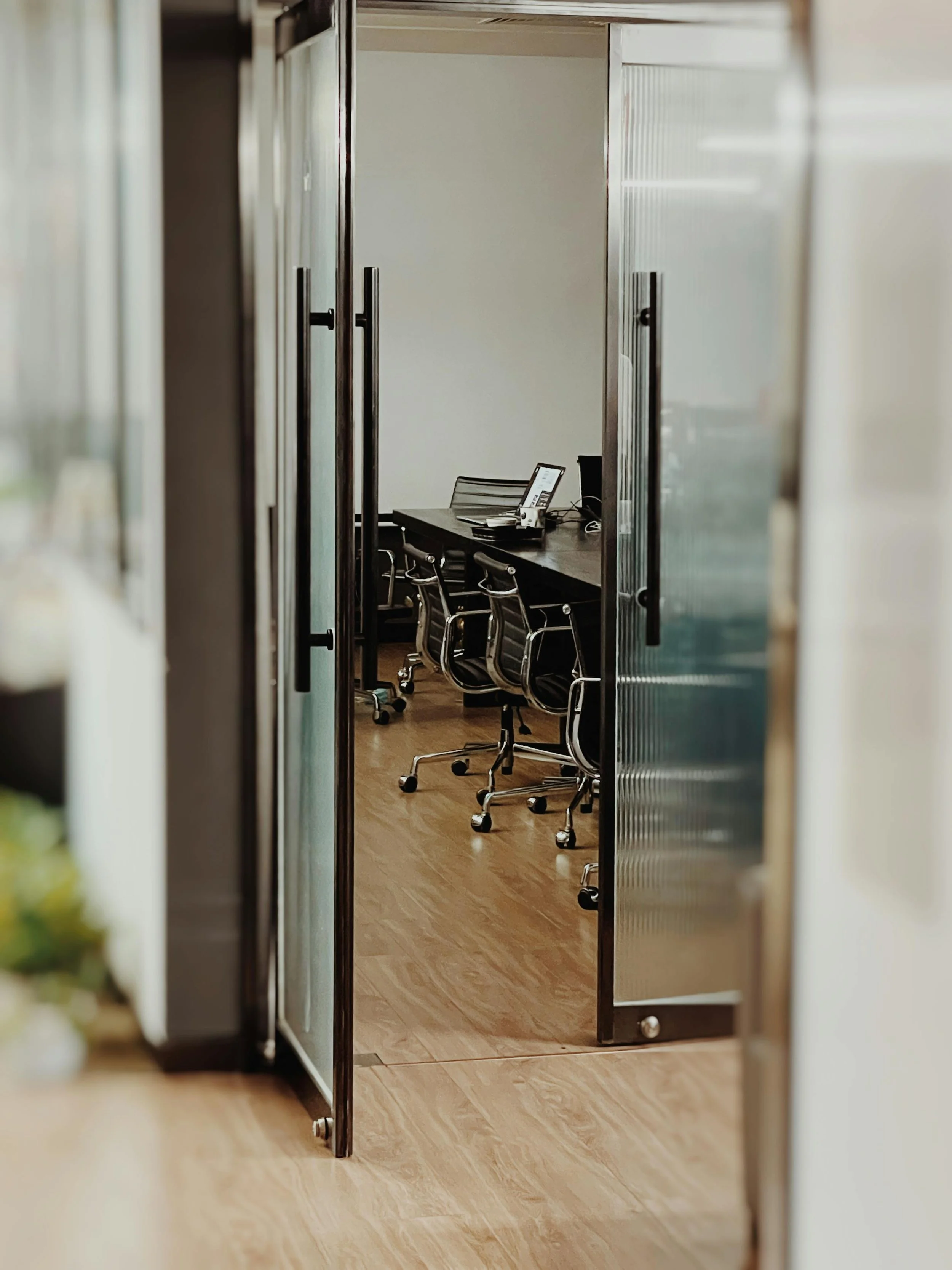 View through partially open glass door into a conference room with black office chairs around a black table, a computer monitor, and a white wall.