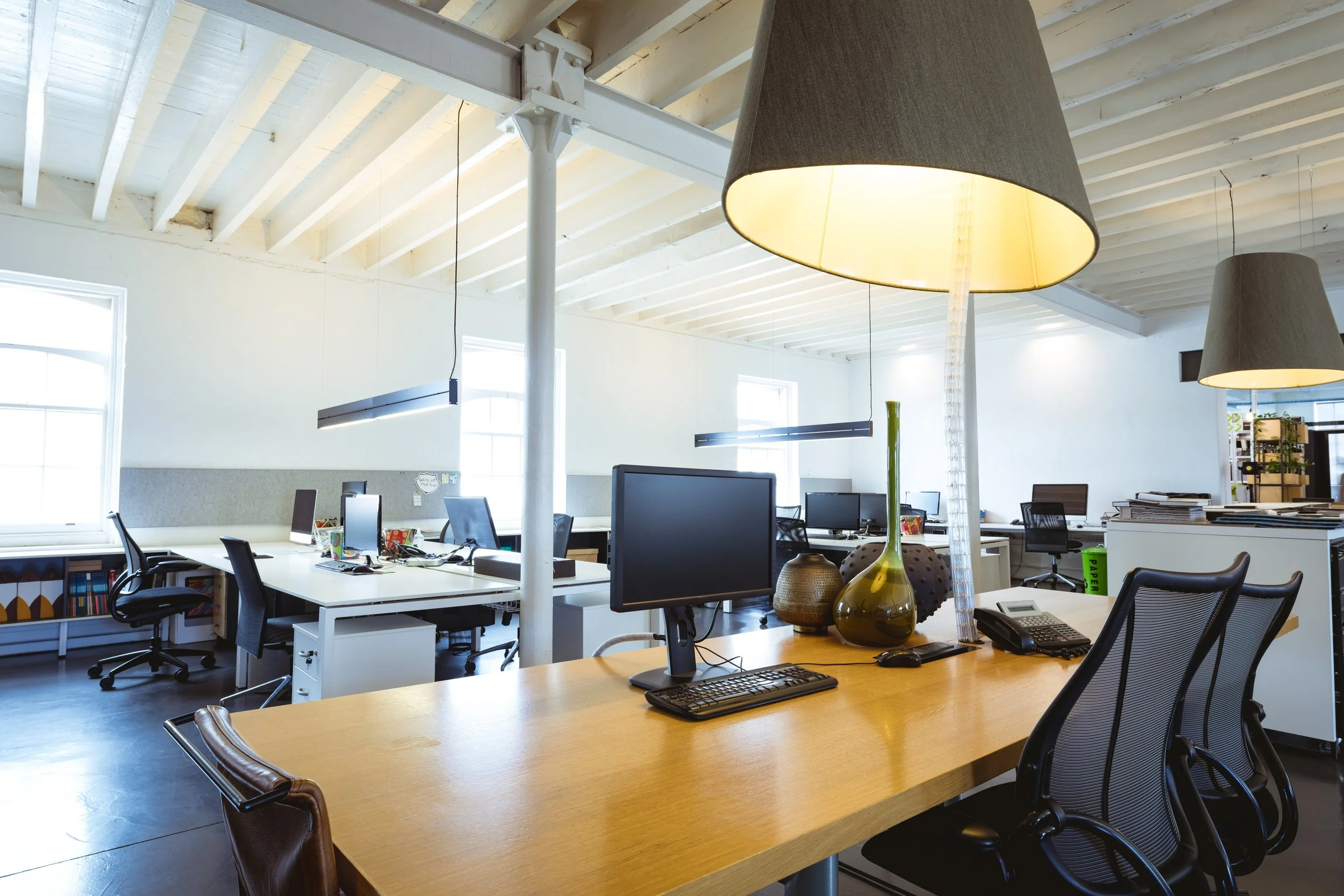 Open-plan office space with white walls, large windows, modern work desks, computer monitors, office chairs, pendant lamps, and decorative vases on a wooden table.