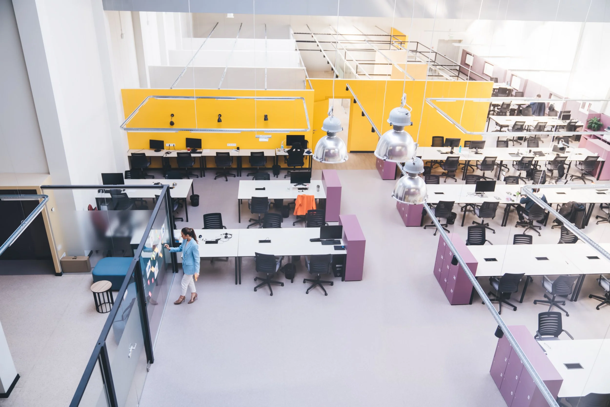 An open-plan modern office with multiple desks and chairs, a woman working with a smartboard, purple filing cabinets, yellow partitions, large windows, and modern lighting fixtures.