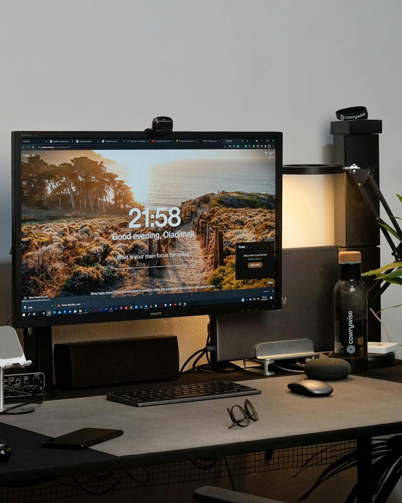 Desk setup with a computer monitor displaying a scenic nature background, a keyboard, mouse, pair of glasses, water bottle labeled CowWwise, and various electronic accessories.