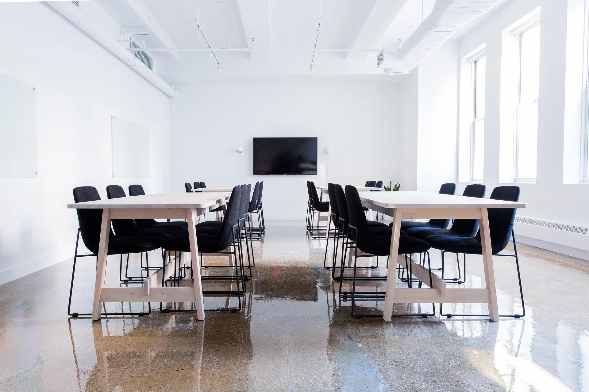 Empty modern conference room with white walls, black chairs, light wooden tables, a mounted flat-screen TV, and large windows.
