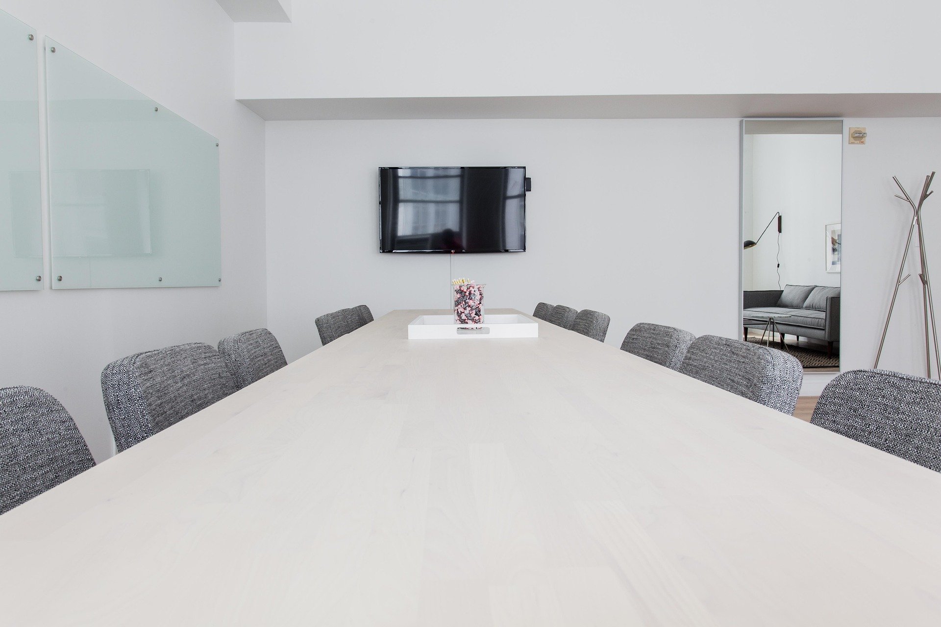 A modern conference room with a large white table, gray upholstered chairs, a wall-mounted TV, a glass board on the wall, and a door leading to a sitting area with a gray sofa and black floor lamp.