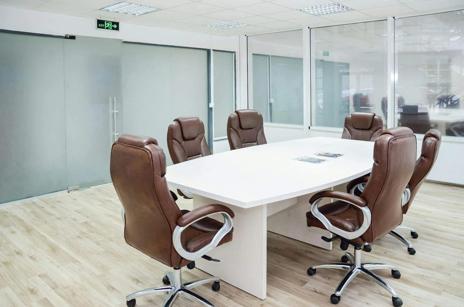 Empty conference room with a white rectangular table surrounded by six brown leather office chairs, glass walls, and a wooden floor.
