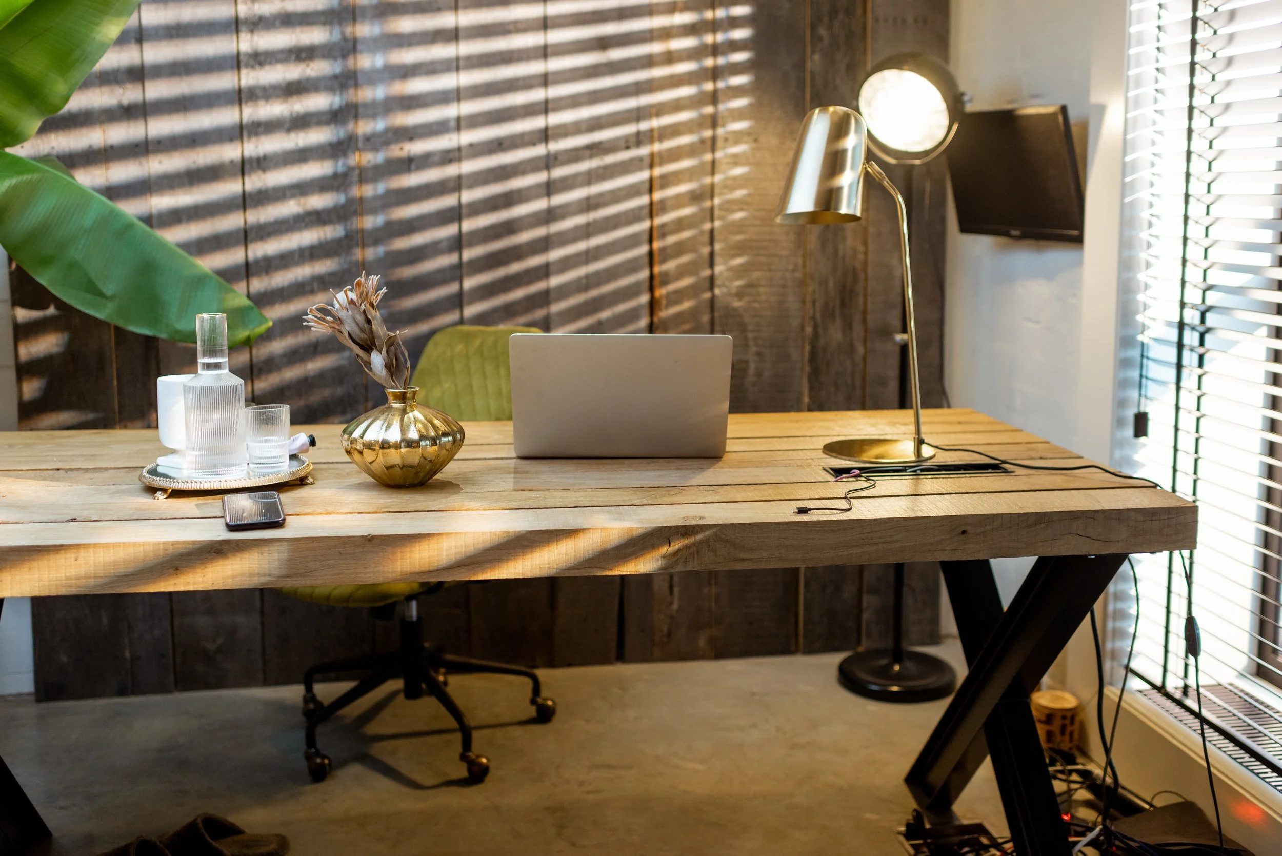 Modern home office with a wooden desk, laptop, glassware, a decorative gold vase, and a brass desk lamp. Sunlight filters through window blinds, casting shadows on the wall. There's a green chair and a TV mounted on the wall.