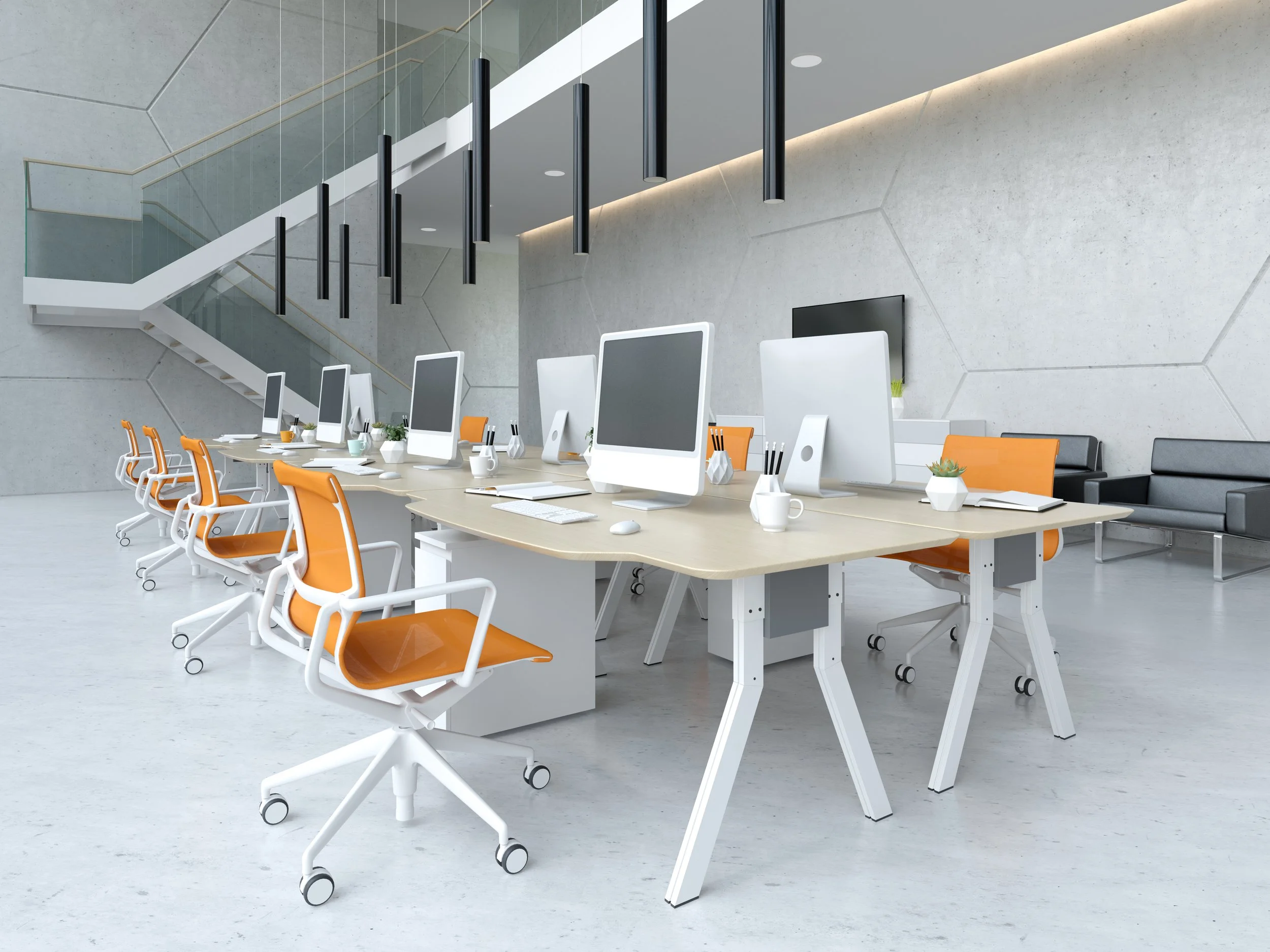 Modern office workspace with a long table, white chairs with orange seats, desktop computers, potted plants, notebooks, and pens, set against a concrete wall with geometric patterns and a staircase in the background.