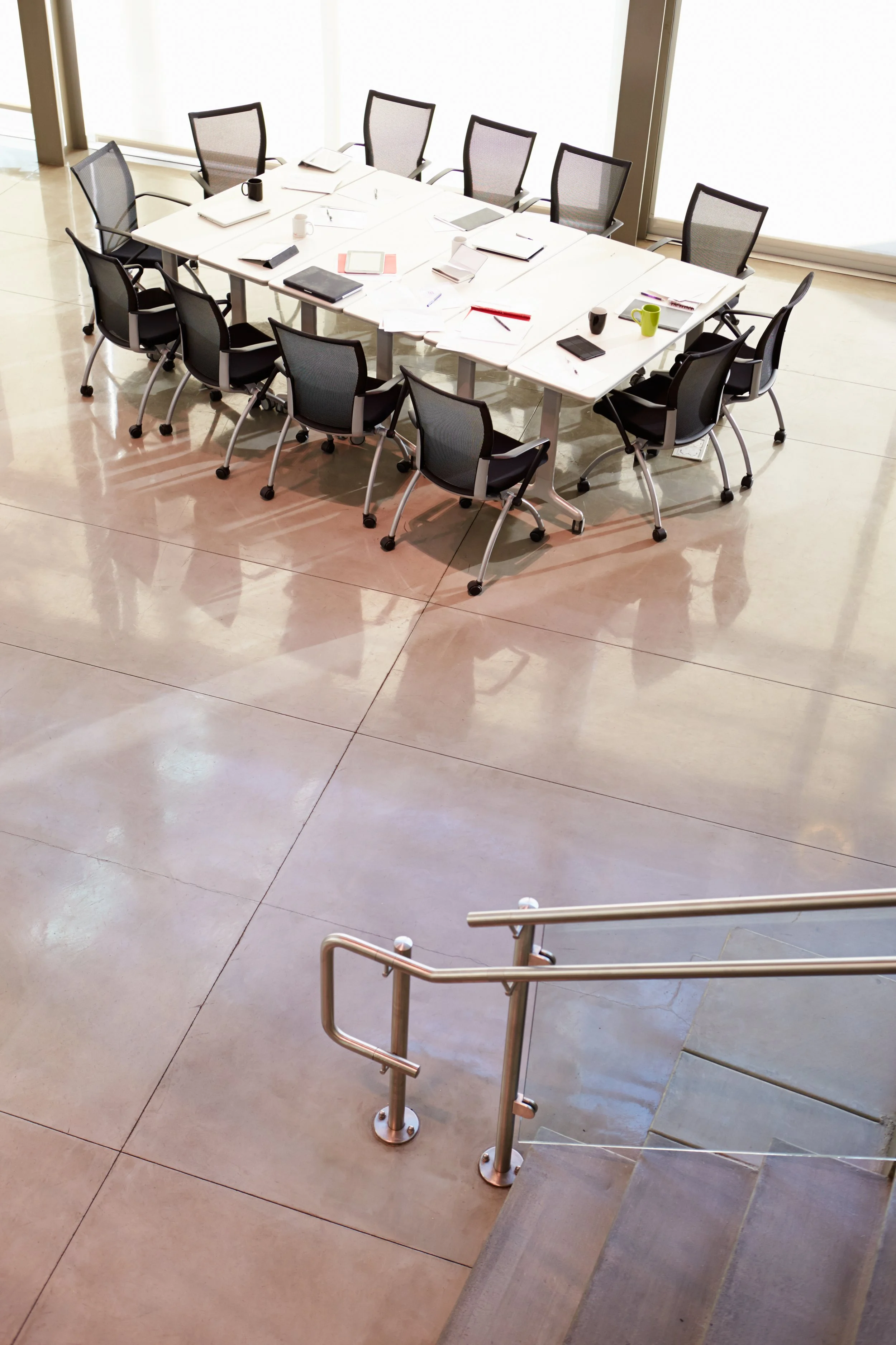 An empty conference room with a large white table surrounded by black mesh office chairs. The table has notebooks, cups, and electronic devices. The room has large windows with frosted glass and a staircase with a metal handrail is visible in the for